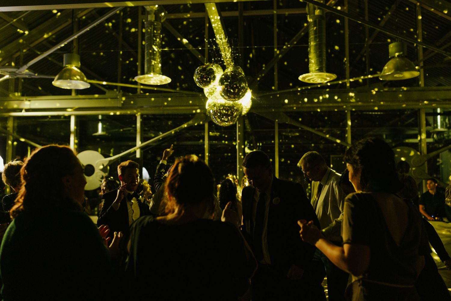 A disco ball illuminates a dark dance floor as party guests celebrate beneath twinkling lights.