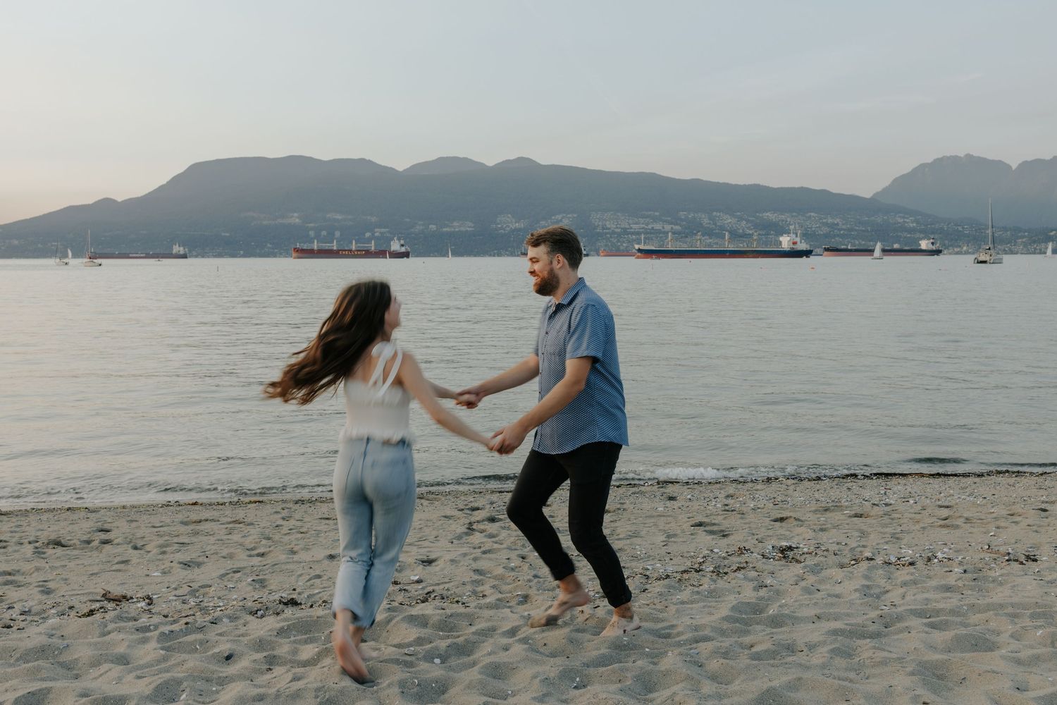 A couple holds hands and dances playfully on a sandy beach with mountains and ocean in the background at dusk.
