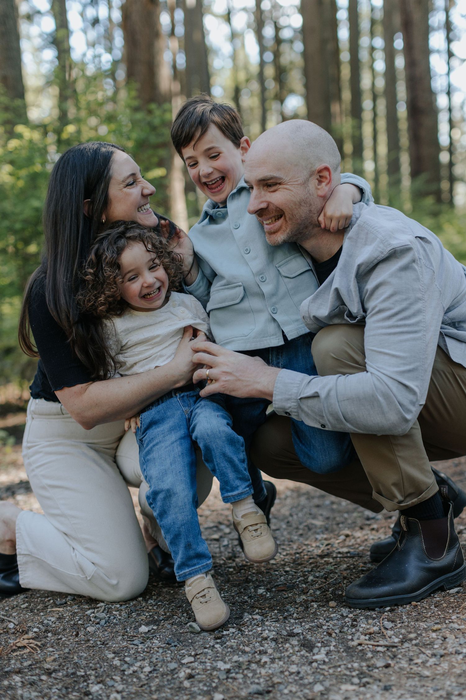 Happy family of four laughing together in a forest, parents crouching with two young children outdoors.