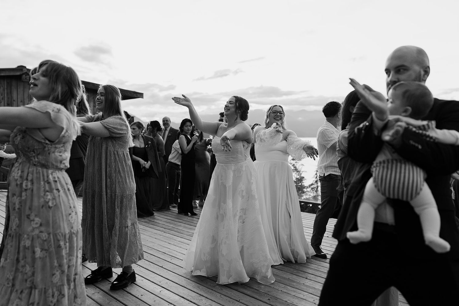 Wedding guests dance together on a wooden deck with mountains in the background during sunset.