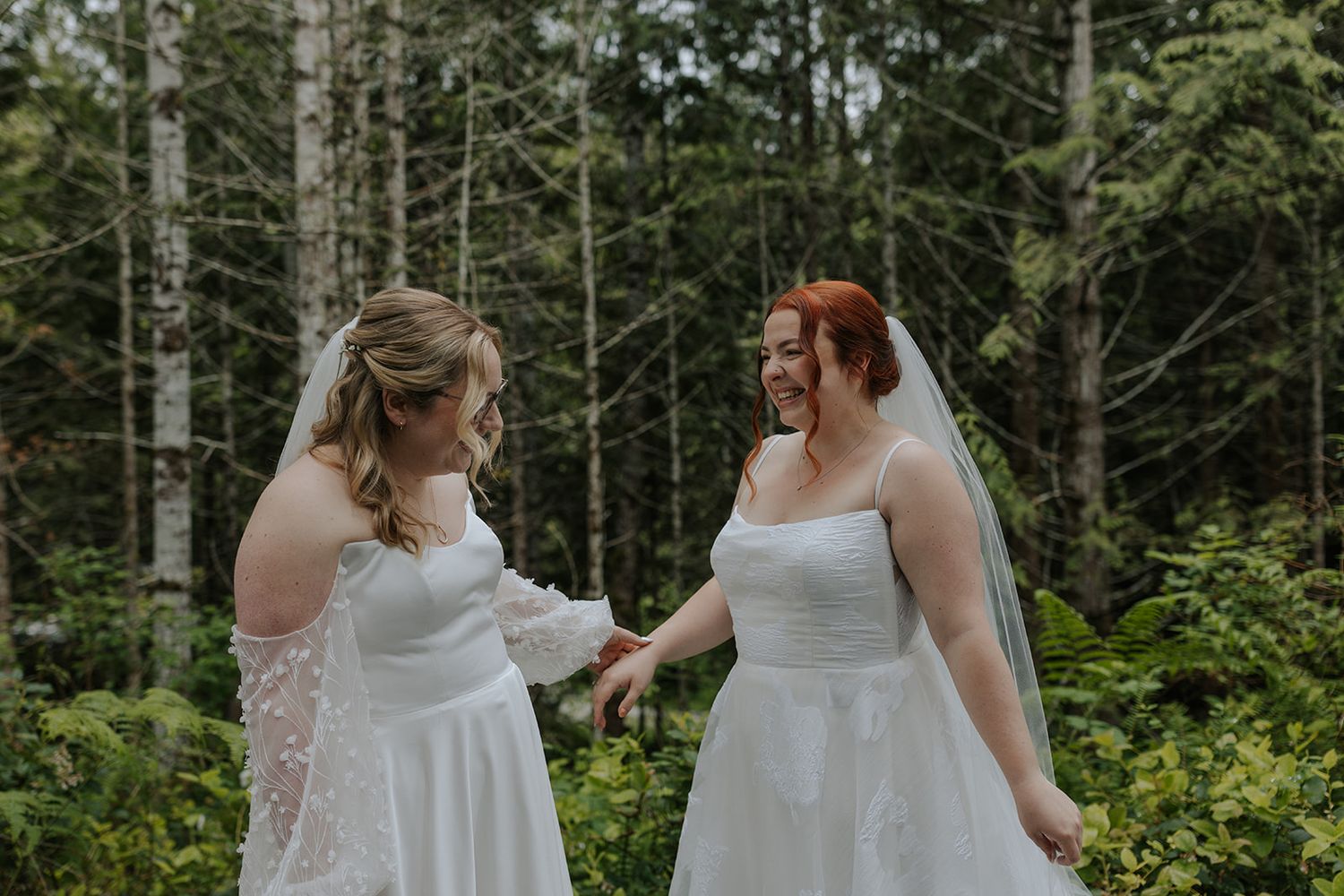 Two brides share a joyful moment together in a forest setting wearing elegant white wedding dresses.