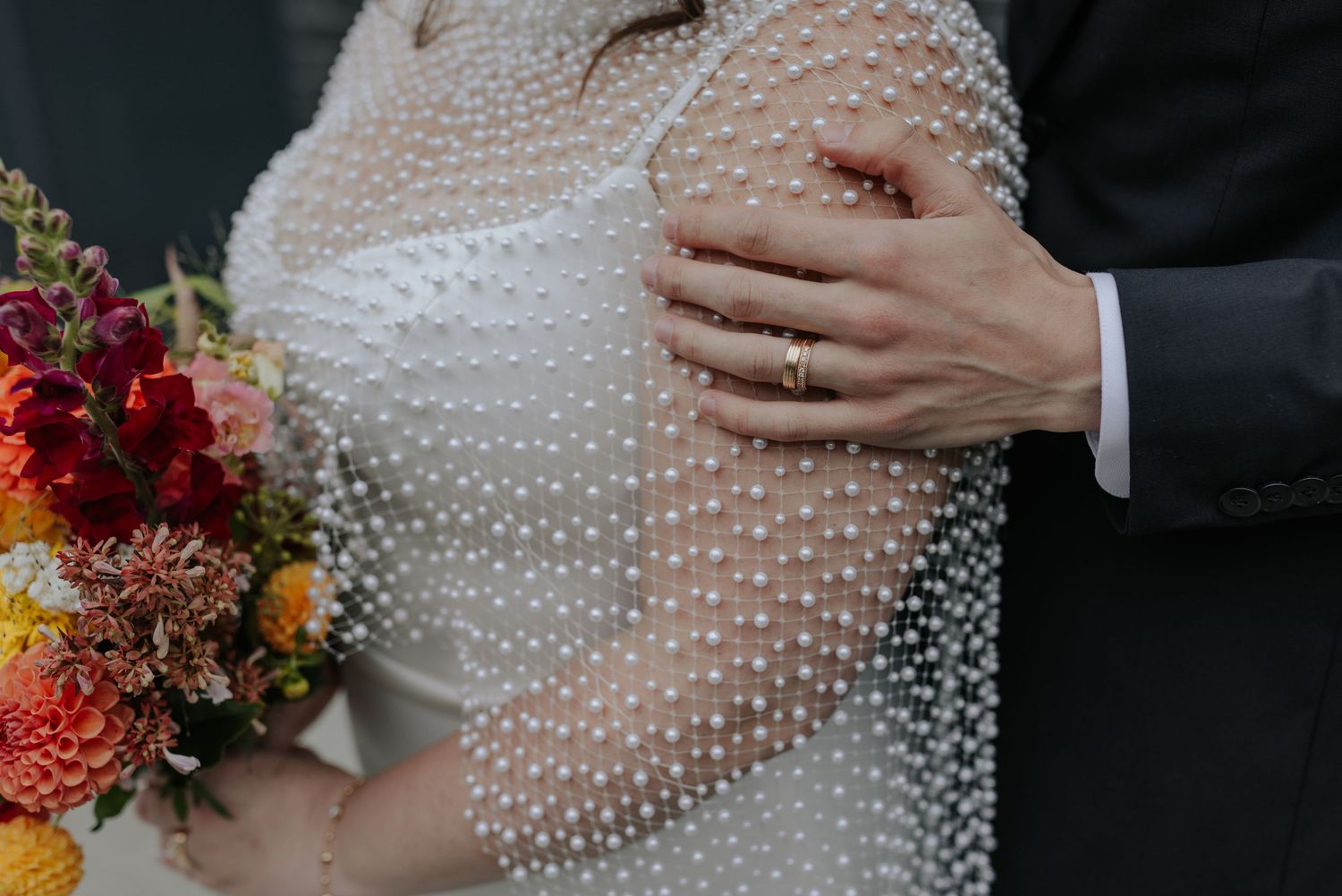 Bride wearing a pearl-beaded dress holds a vibrant autumn-colored bouquet against a modern architectural backdrop.