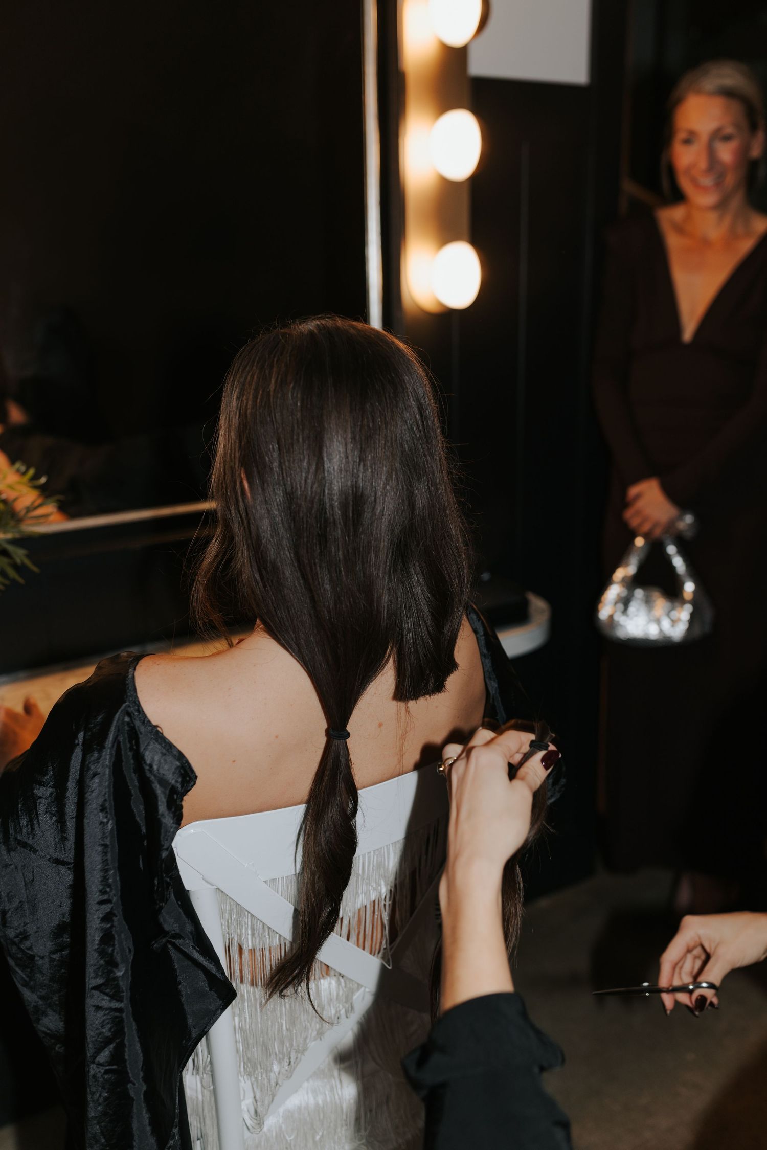 A woman in a white dress gets ready backstage with vanity lights in the background.