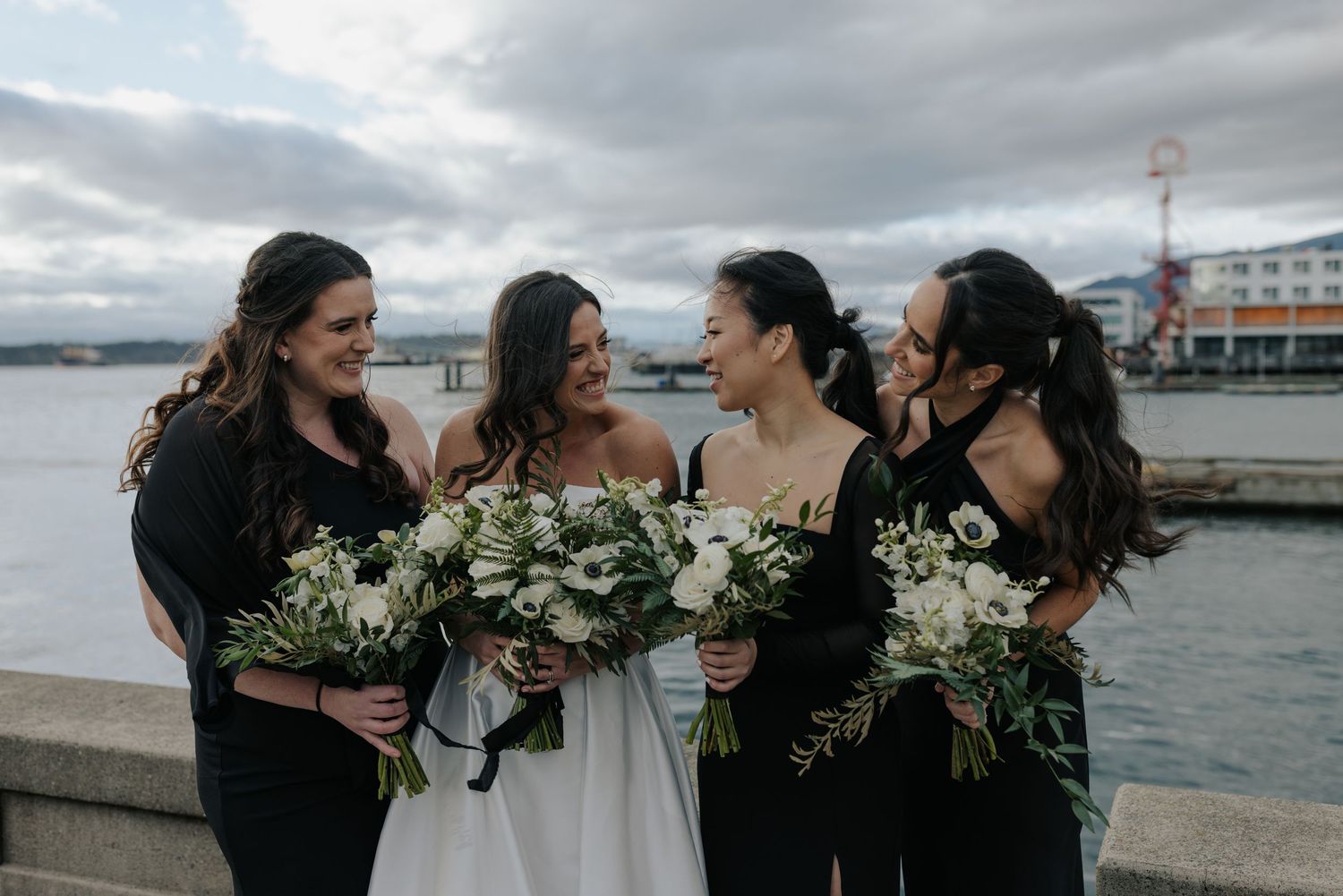 A group of bridesmaids in black dresses share laughter and smiles while holding white floral bouquets by the waterfront.