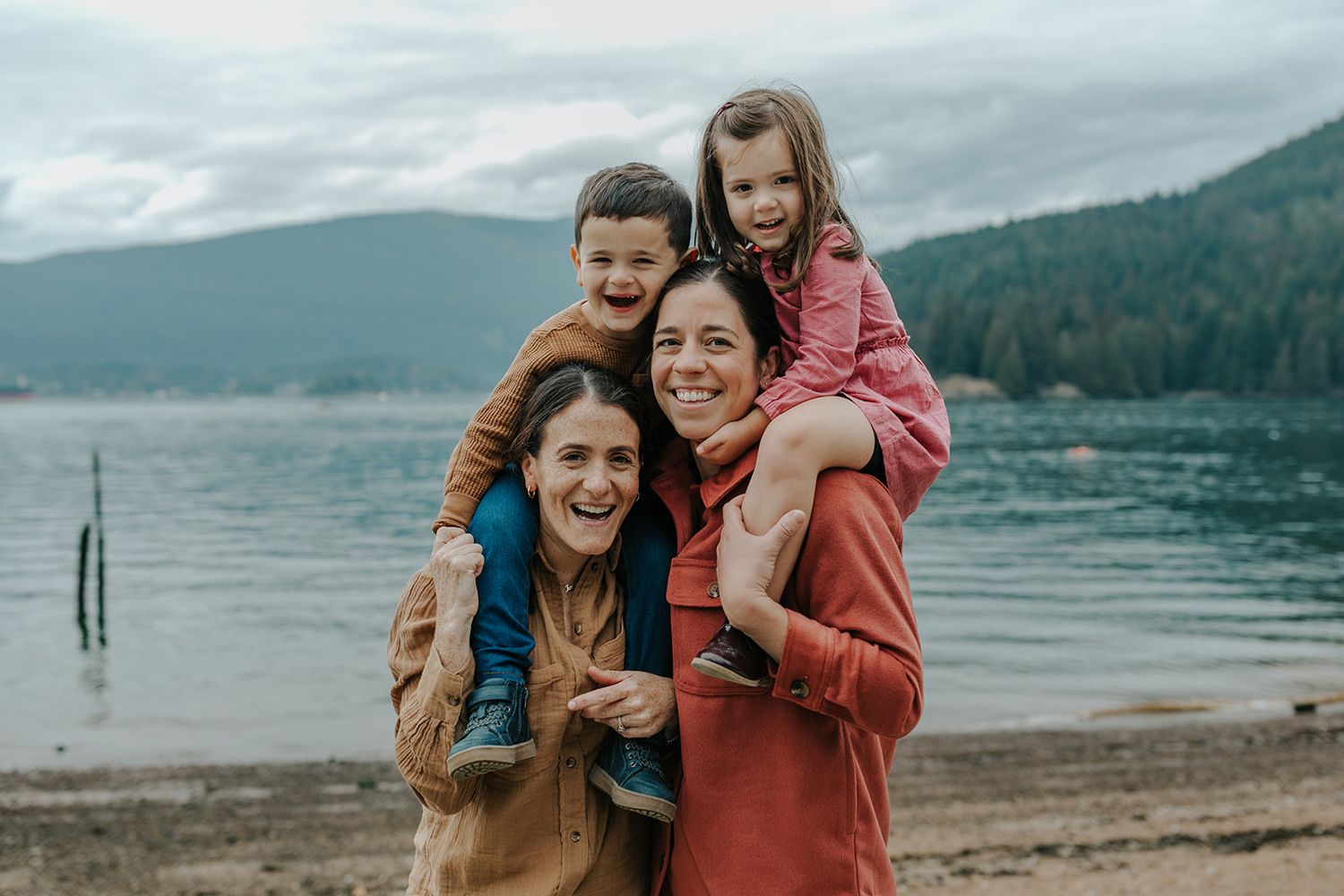 Two women smile on a lakeside beach, each giving a young child a piggyback ride, with mountains in the background.