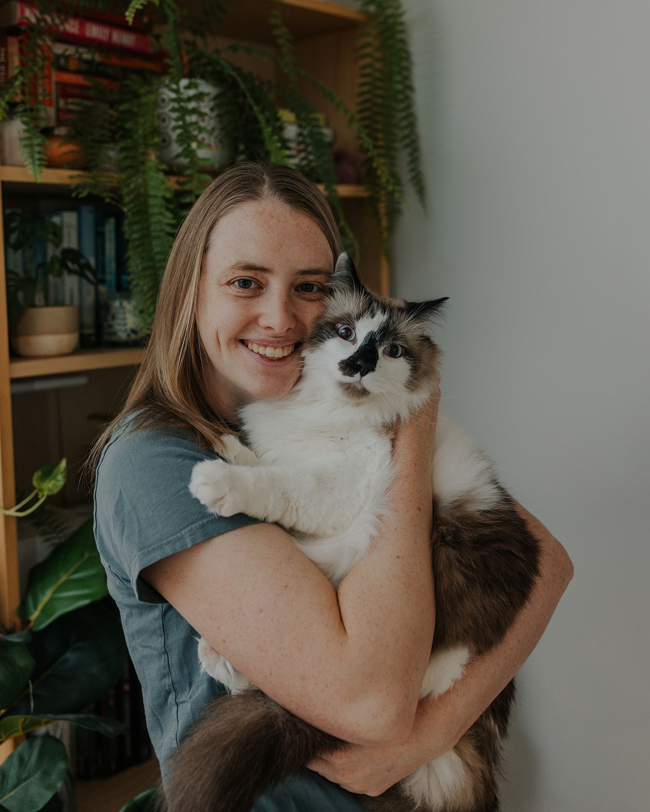 Smiling woman holding a fluffy ragdoll cat indoors with bookshelves and green plants in background.