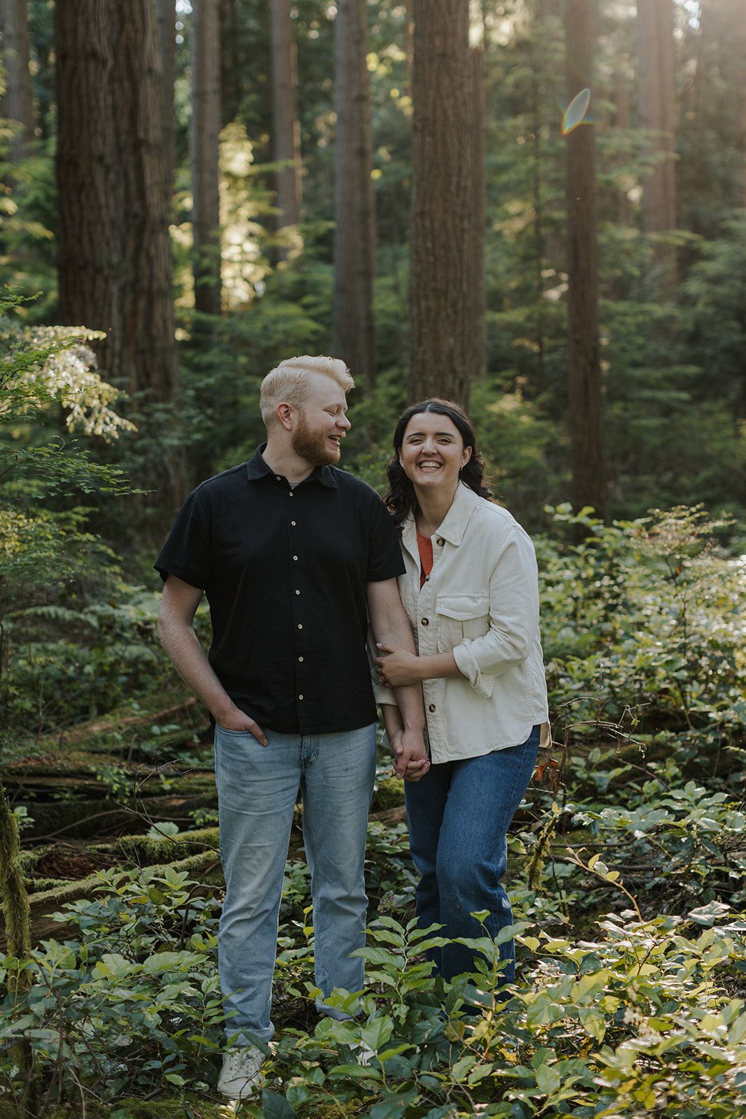 A smiling couple stands together in a lush green forest, the woman in a white jacket holding the man's arm.