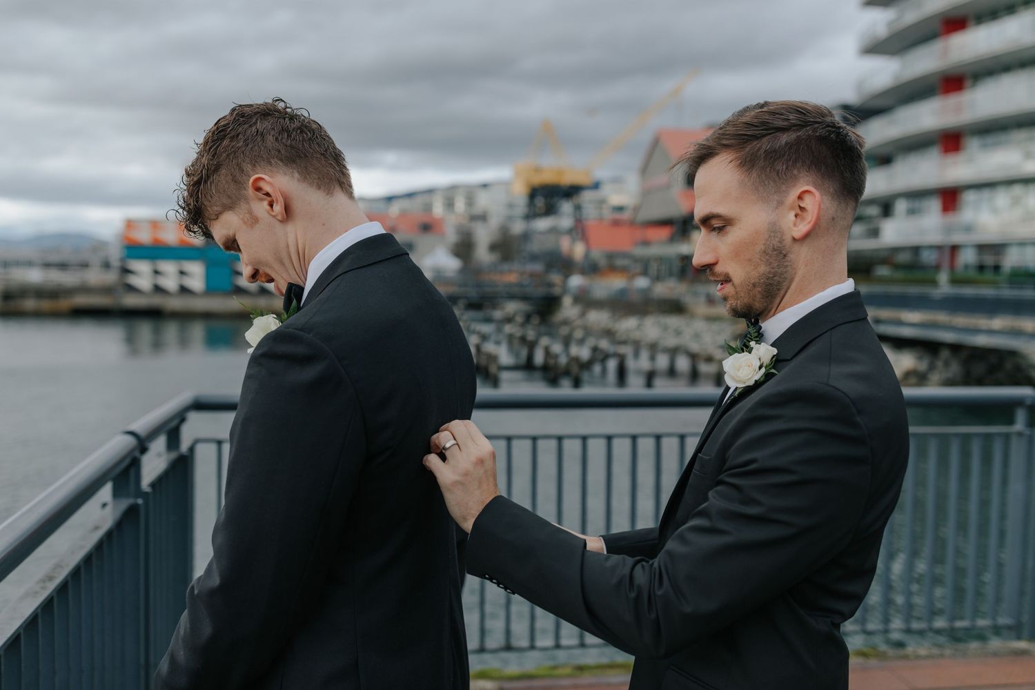 Two people in black suits stand on a waterfront deck with buildings and boats visible in the background.