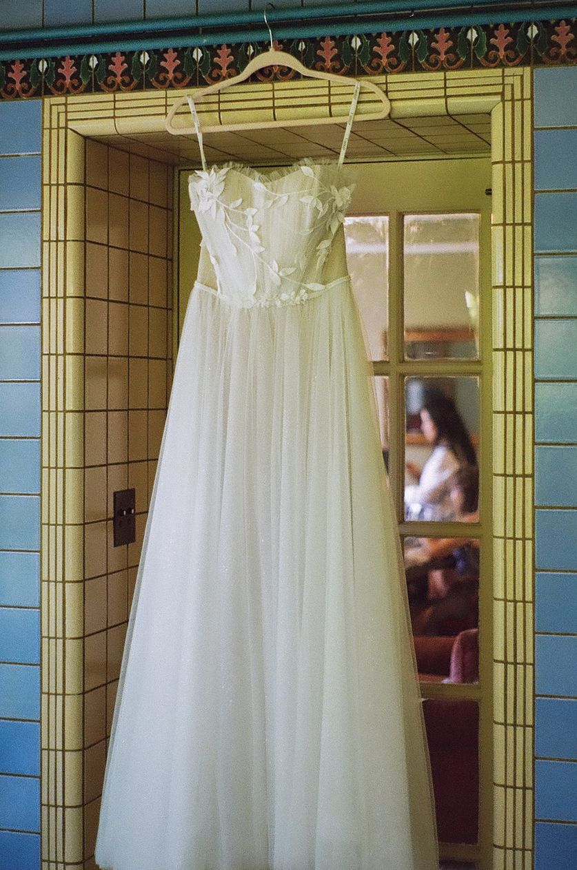White wedding dress hanging in a vintage bathroom with blue tiled walls and ornate wallpaper.
