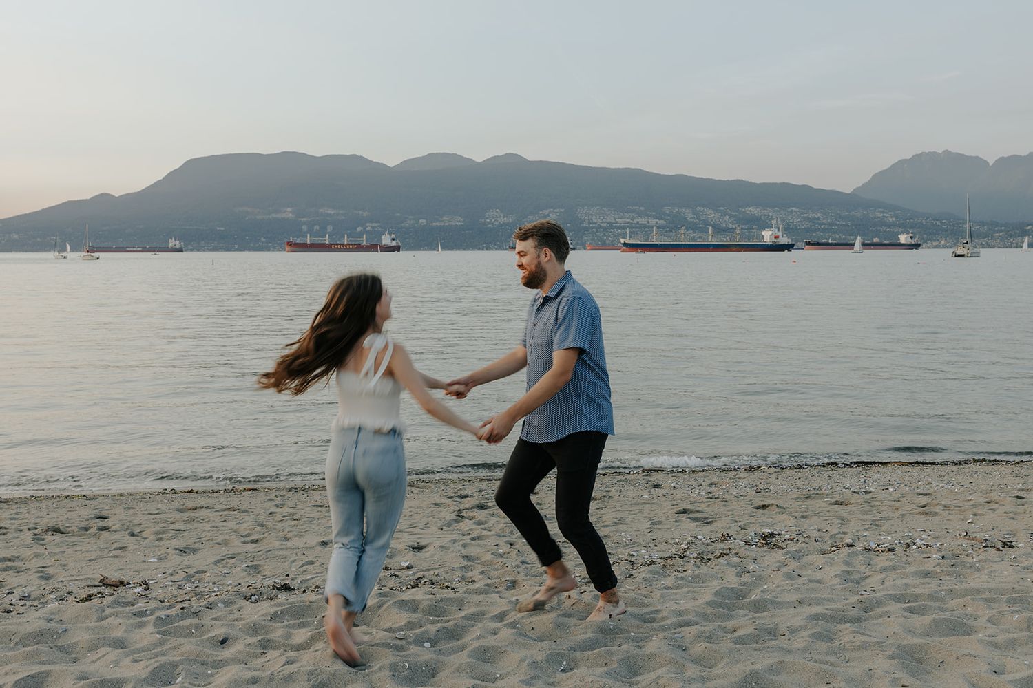 A playful couple dances and spins together on a beach with mountains in the background.