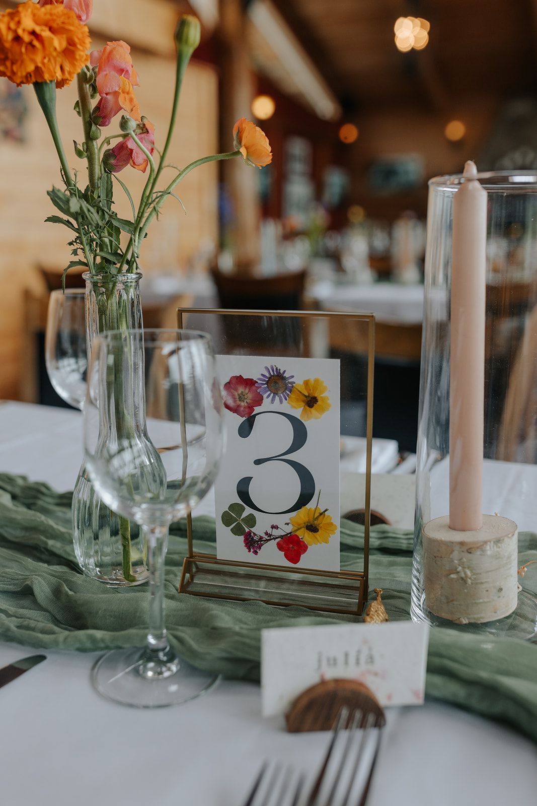 Wedding table decor featuring a floral table number card, candle holder and orange flower arrangement.