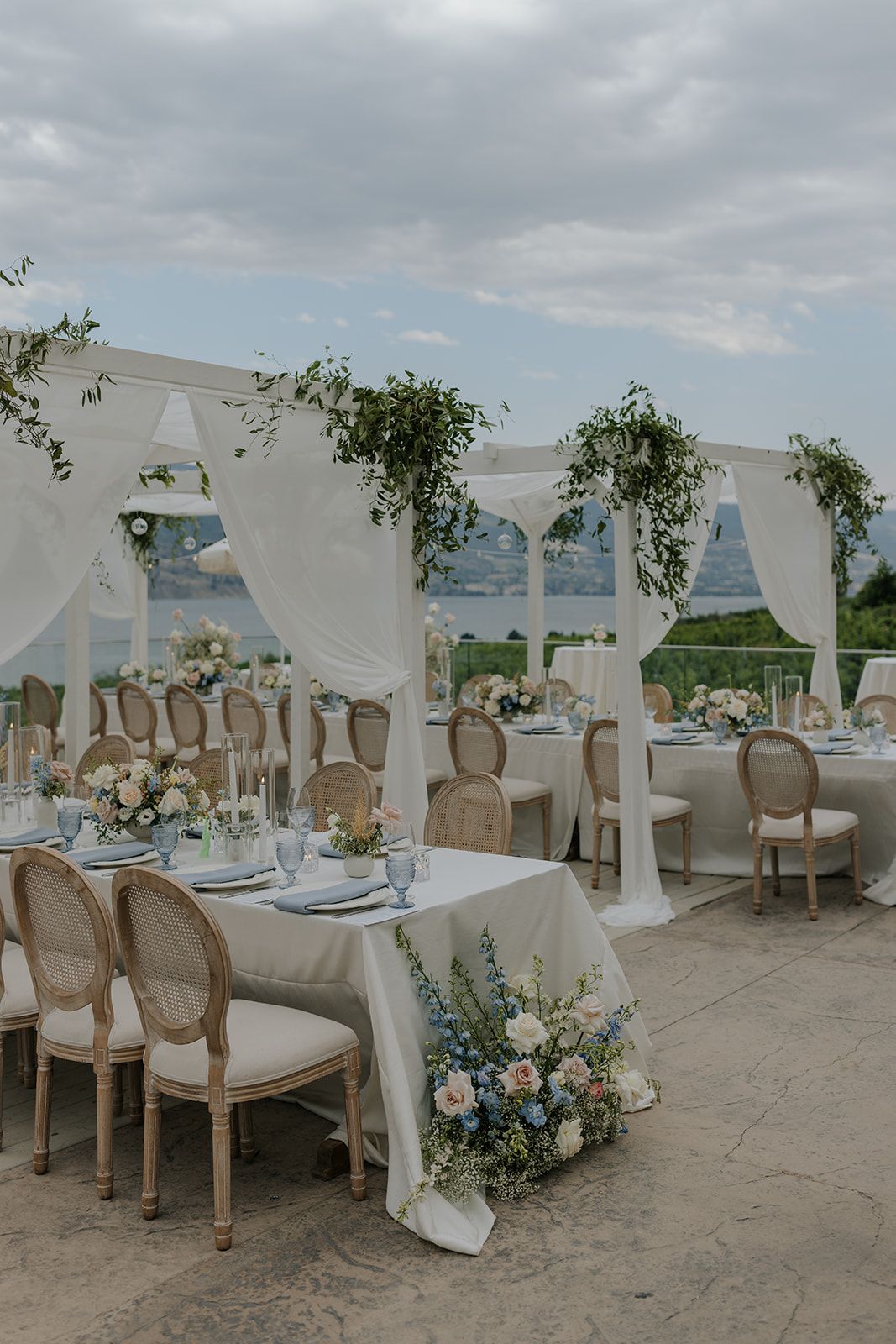 Elegant outdoor wedding reception setup with white drapes, beige chairs and floral centerpieces under cloudy skies.