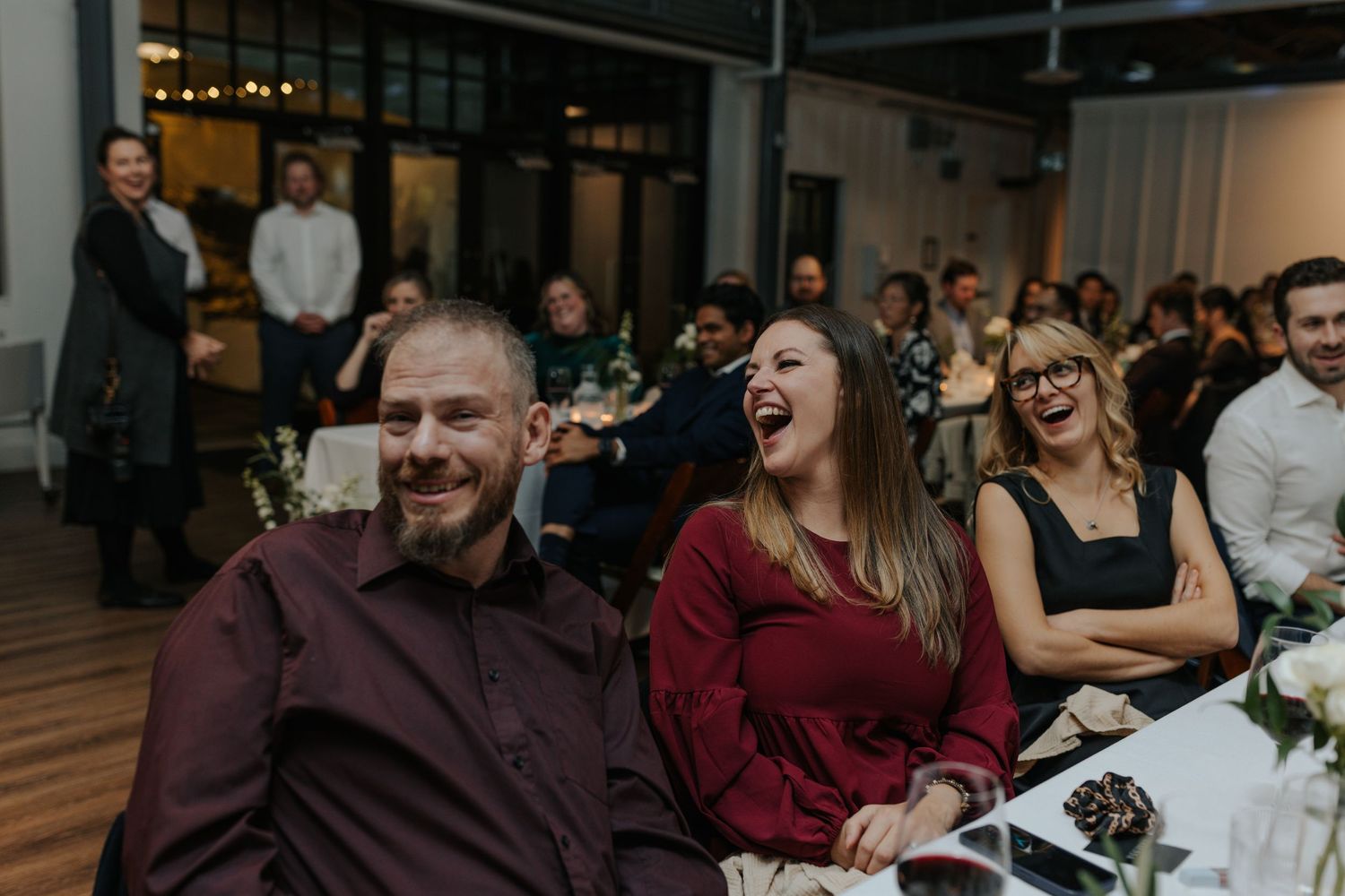 Wedding guests react with laughter during speeches at an indoor reception venue.