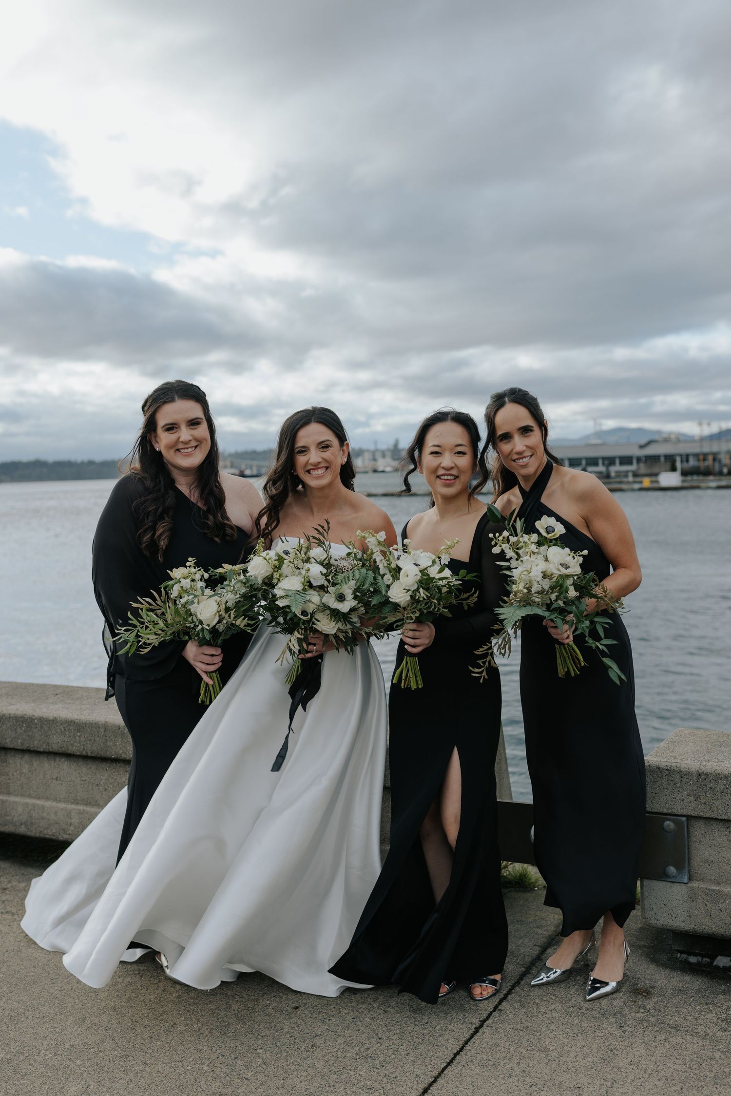 Wedding party in black dresses and white gown pose together along waterfront under cloudy skies.