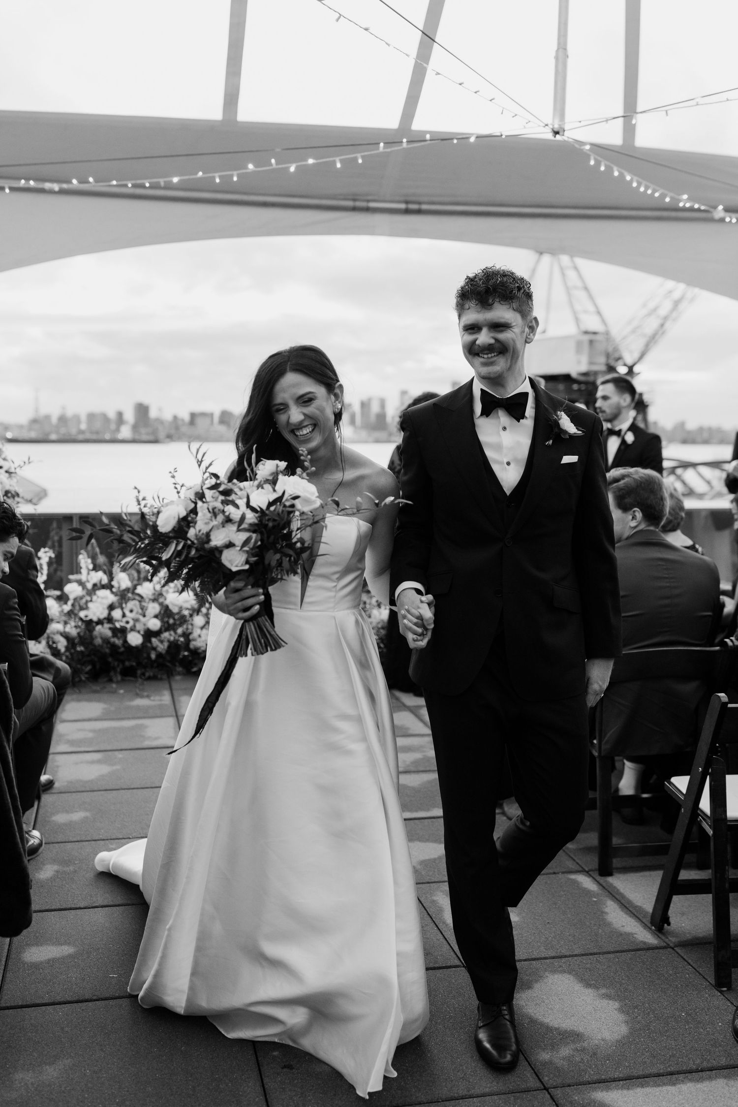 A couple celebrates their wedding day under string lights at an outdoor waterfront venue.