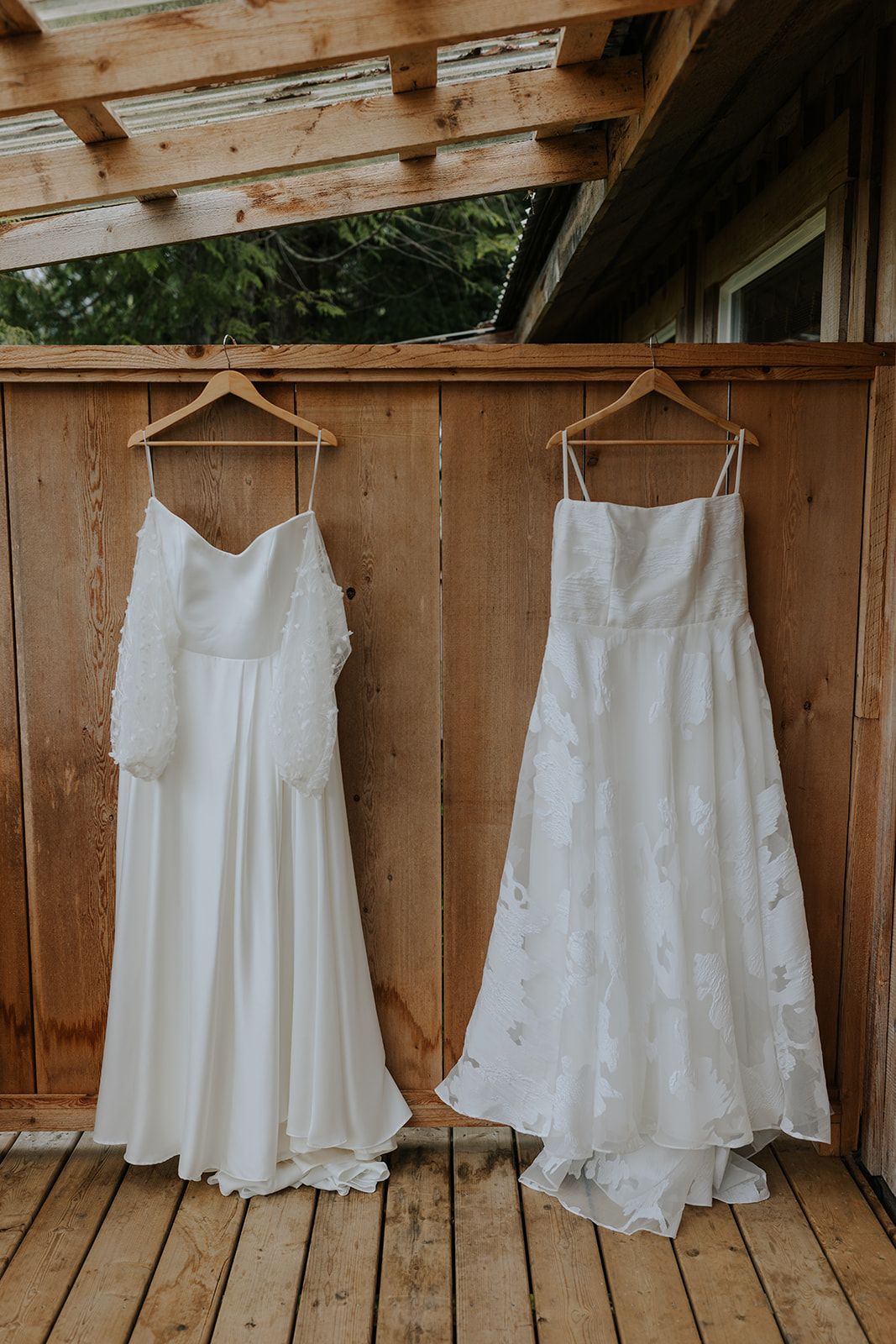 Two elegant white wedding dresses hanging on wooden hangers against a rustic wooden wall.
