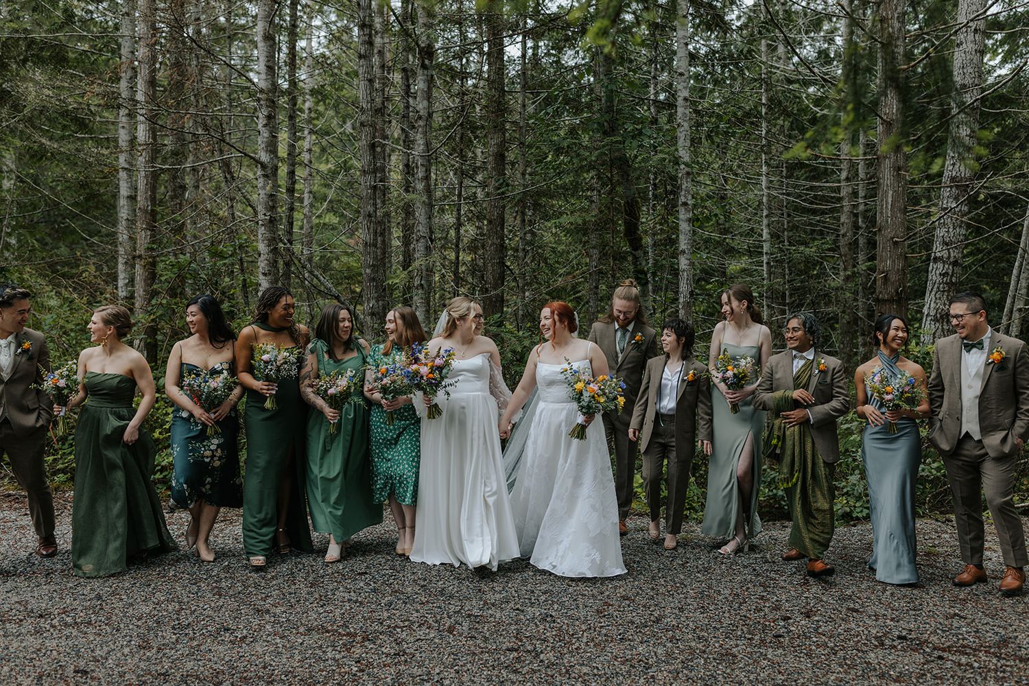 Wedding party members lined up on forest path wearing mix of green and white dresses.