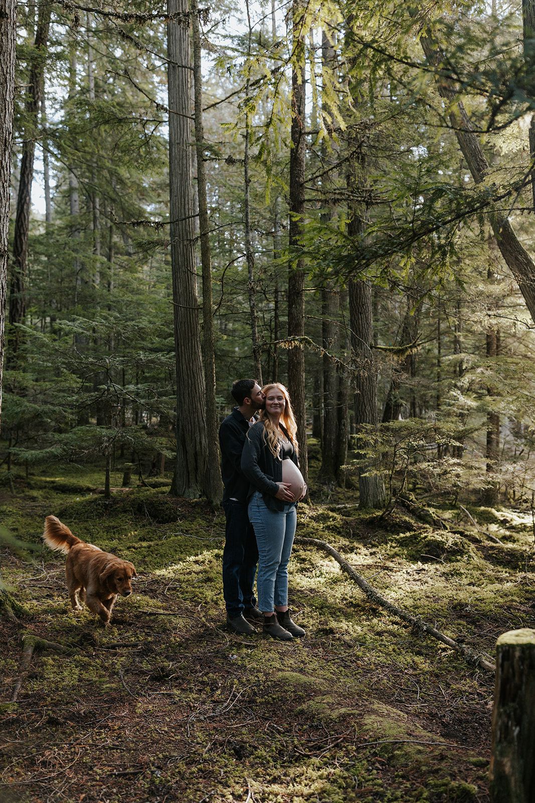 Couple embracing in a forest with their dog walking nearby.