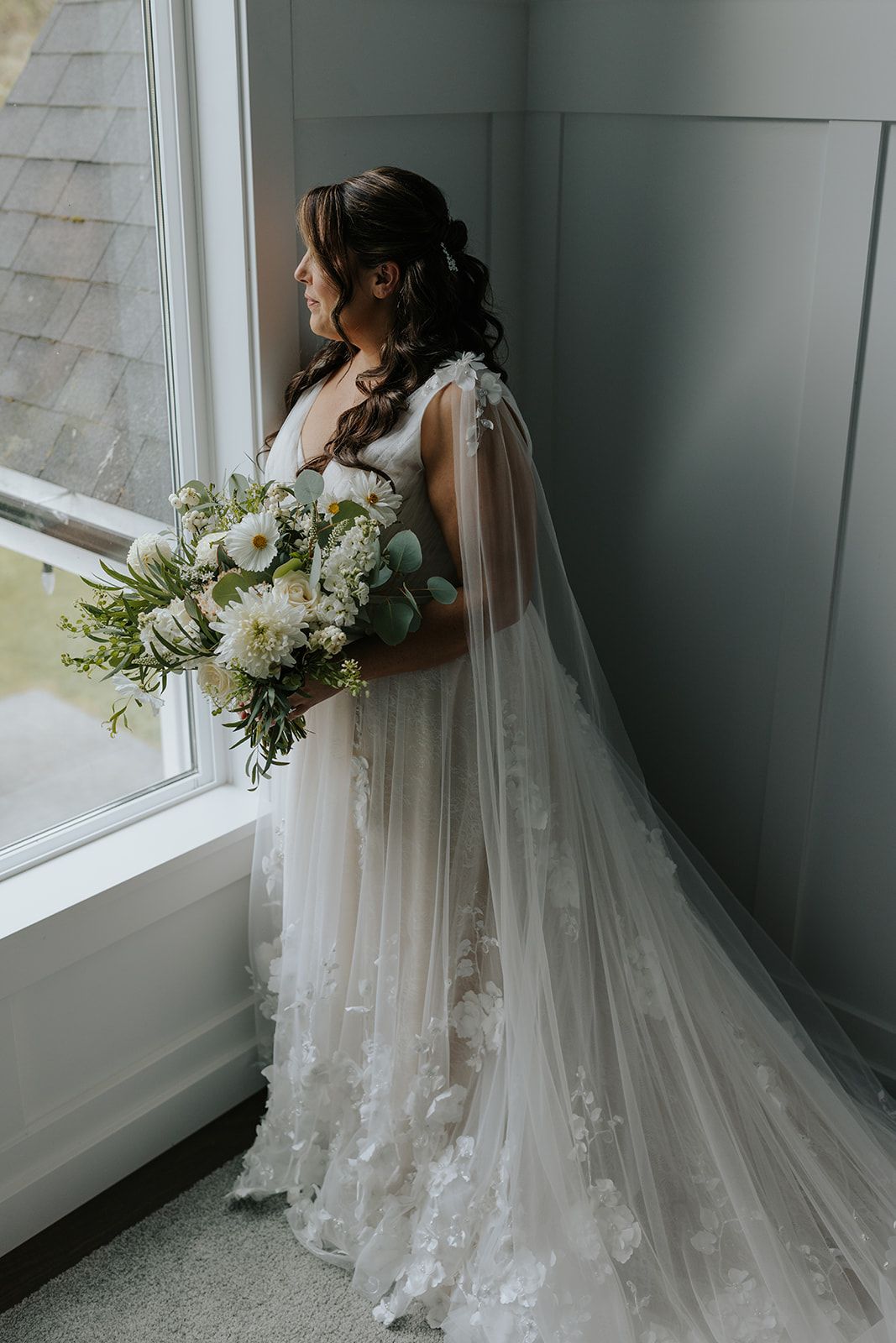 A bride in a flowing lace gown with veil stands by a window holding a white bouquet.