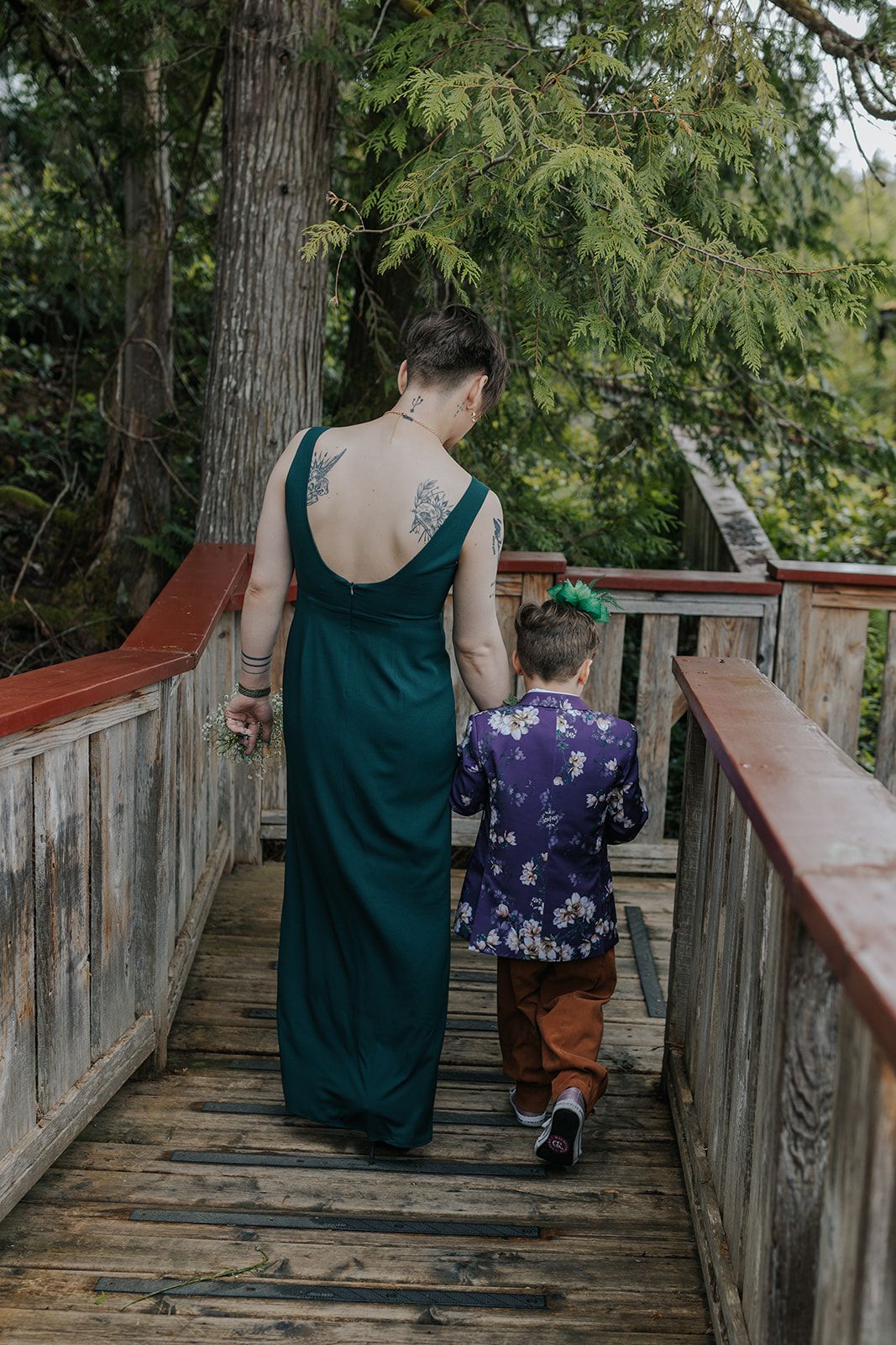 Person in green dress walks with child on wooden boardwalk surrounded by evergreen trees.