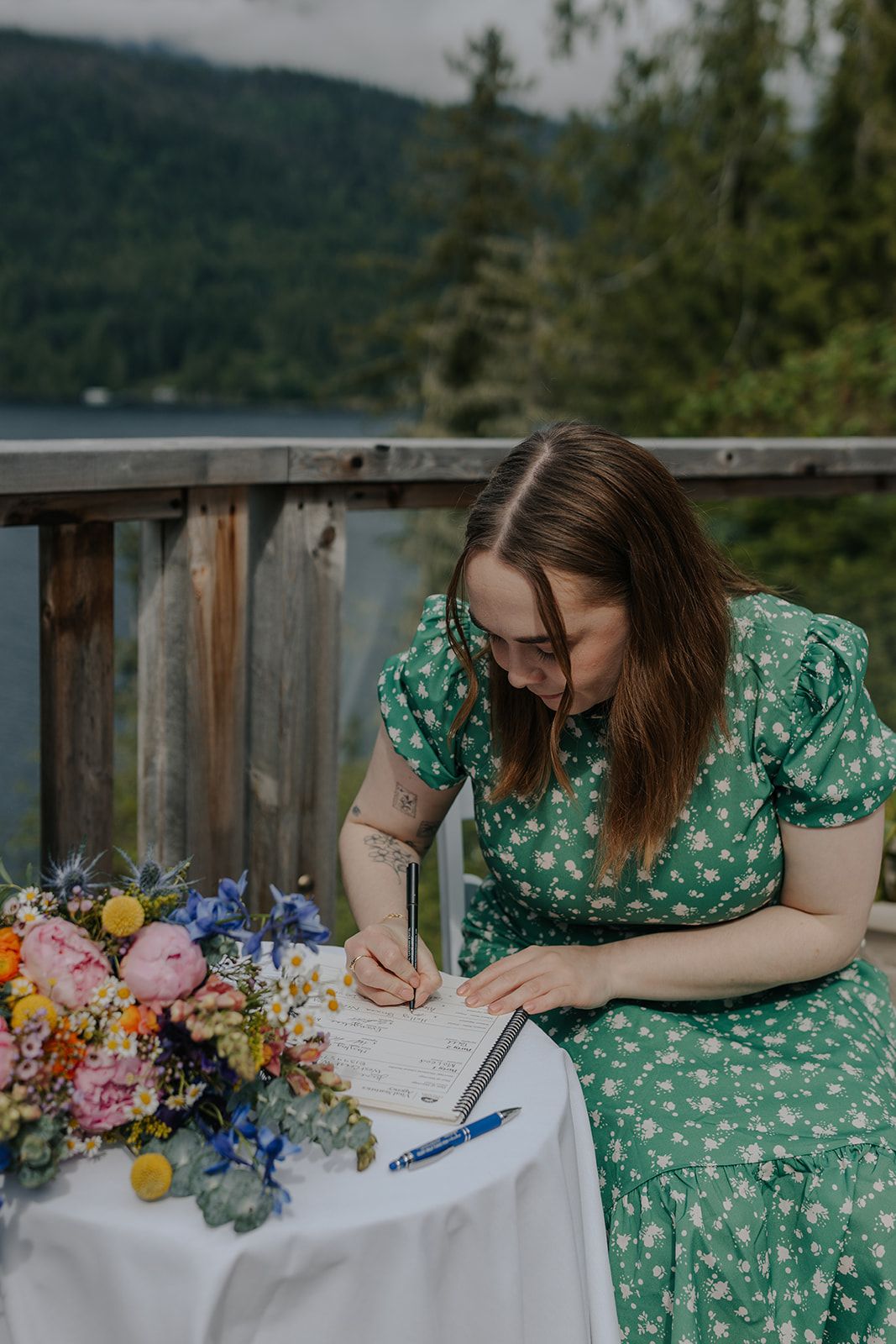 Someone in a green floral dress signs a document next to colorful flowers on an outdoor deck.