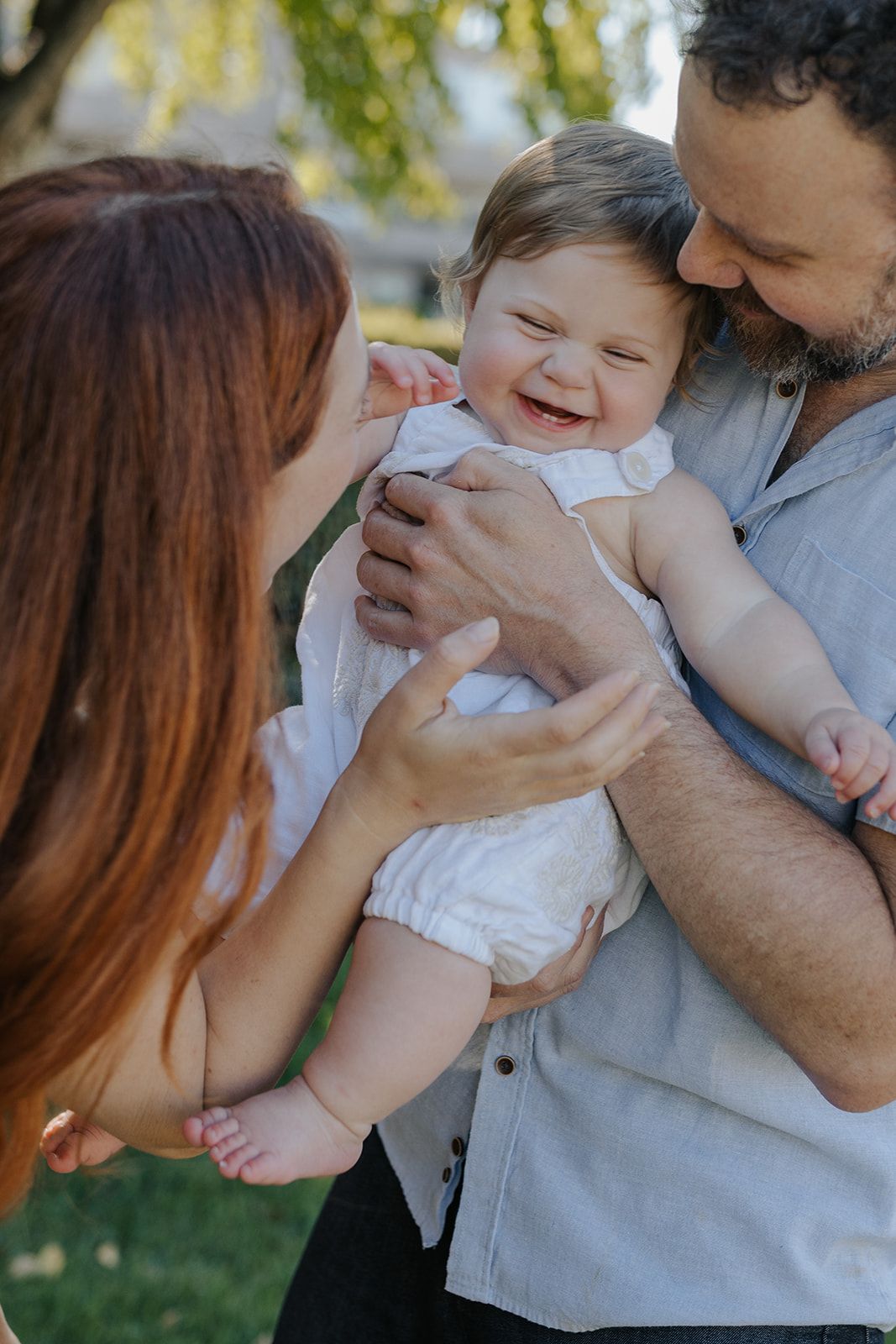 A baby in a white outfit smiles joyfully while being held by parents outdoors on a sunny day.