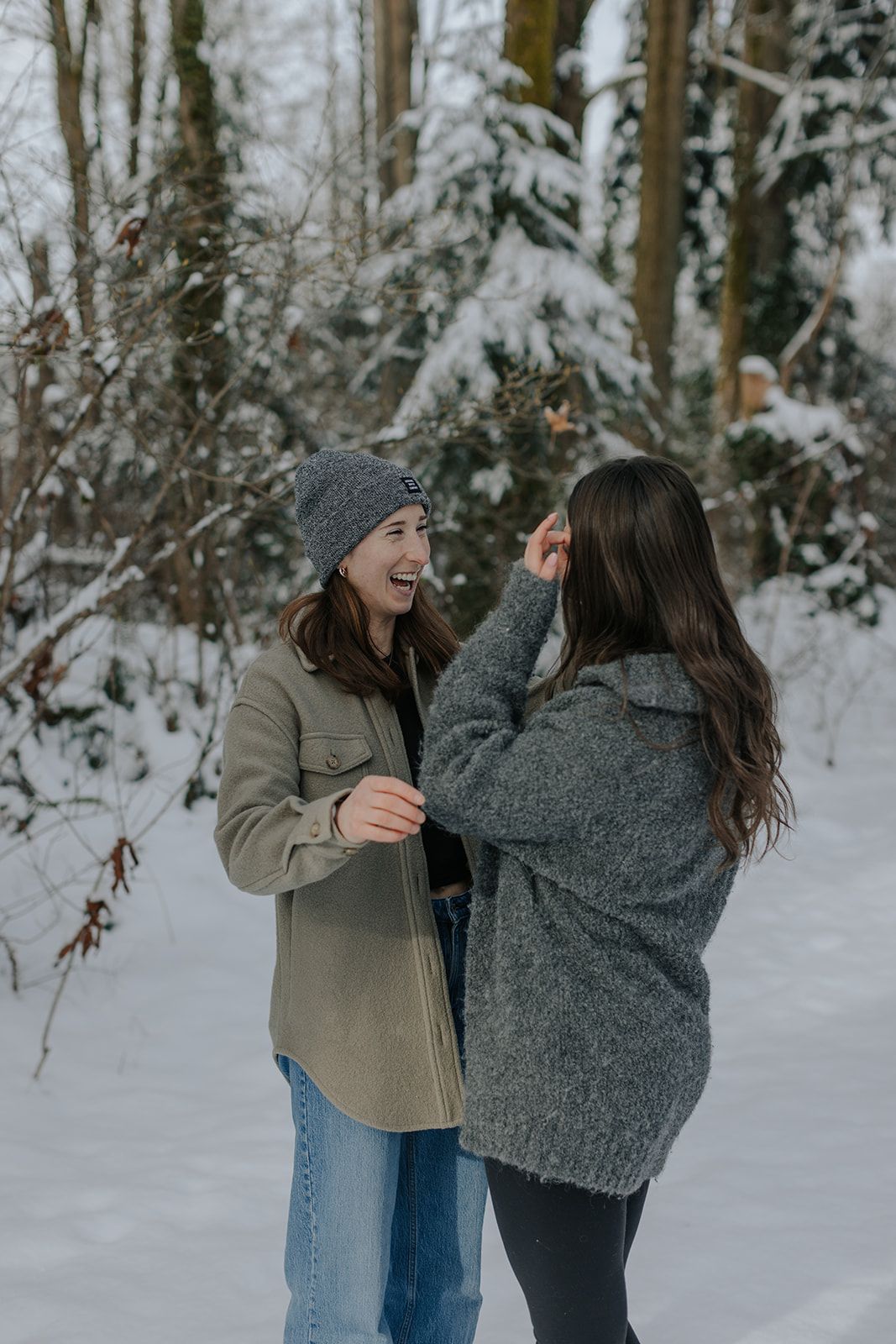 Two women laughing together in a snowy winter forest, wearing cozy jackets and a gray beanie hat.