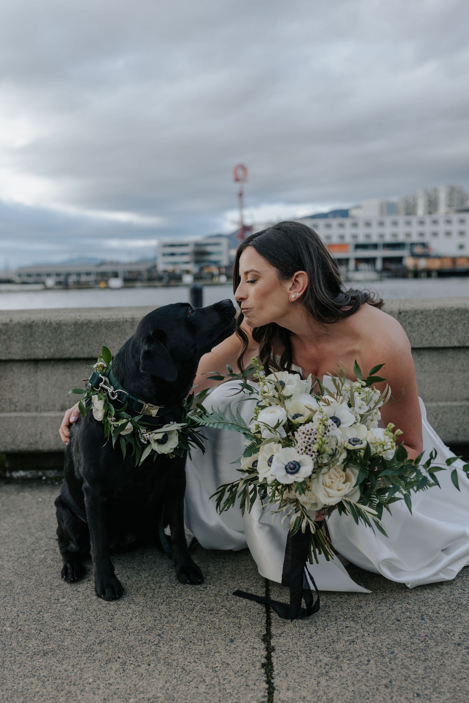 Black dog with floral collar lies on ground next to person in wedding dress during photoshoot.
