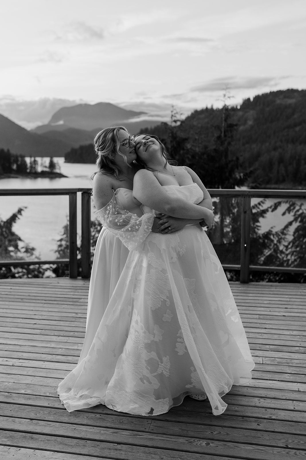 Wedding party members in white dresses sit close together on a wooden deck at sunset.