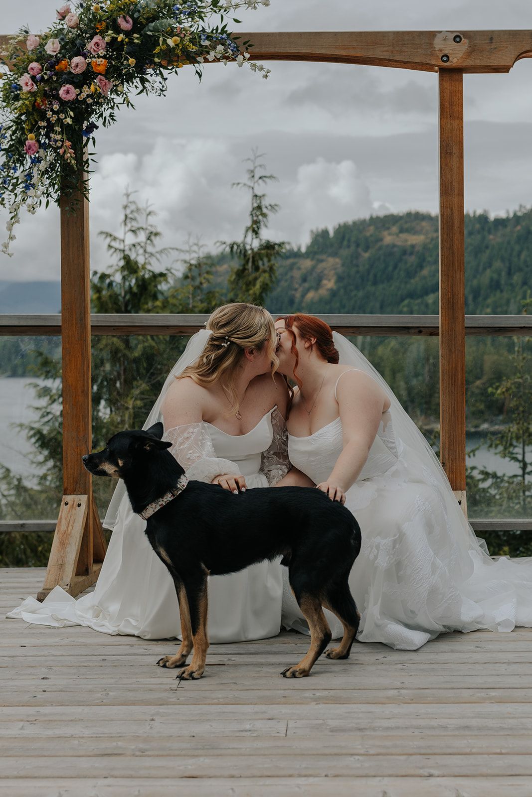 Two dogs join the brides on a wooden deck during wedding photos with mountain scenery.
