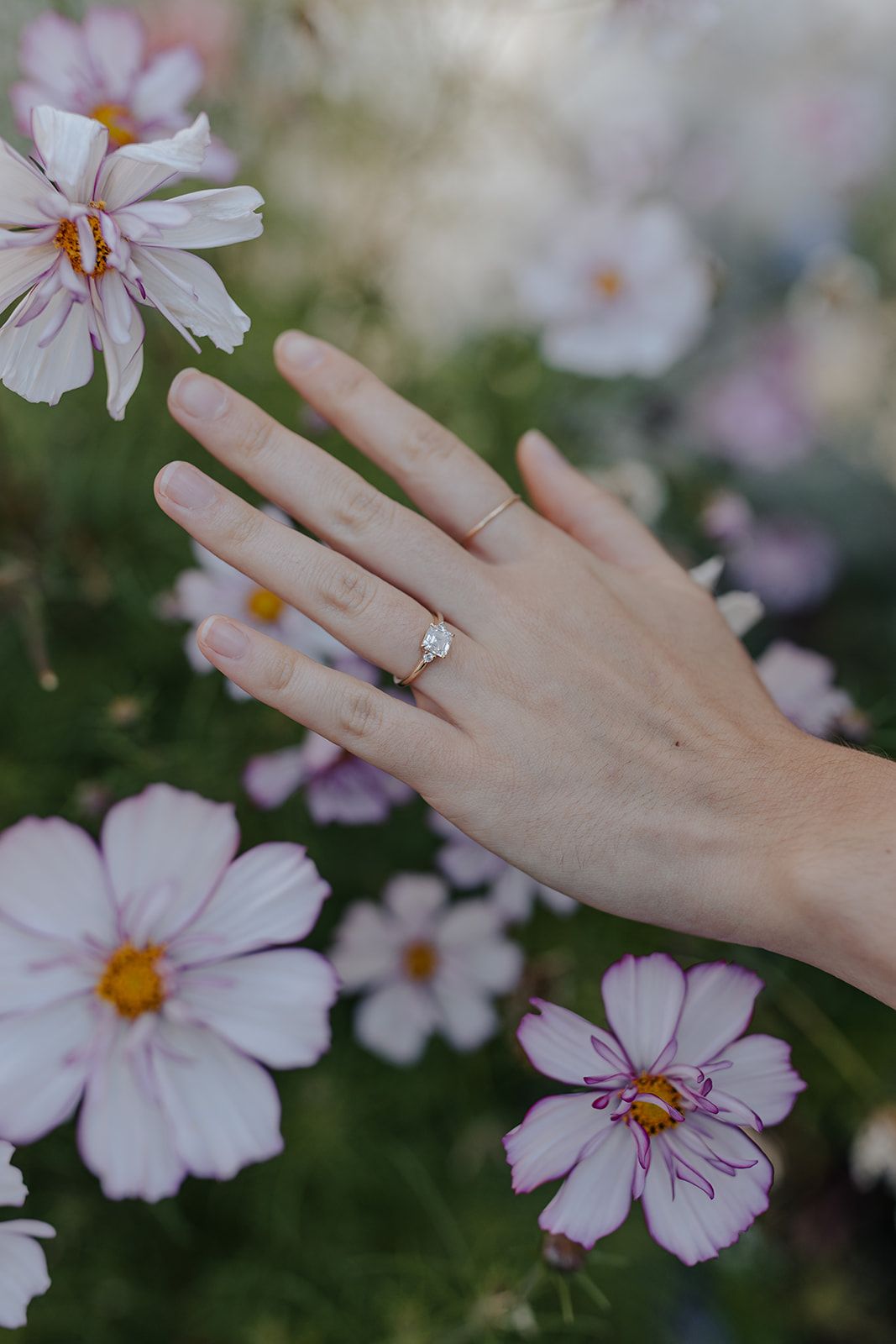 Delicate hand with engagement ring against pink cosmos flowers in a garden setting.