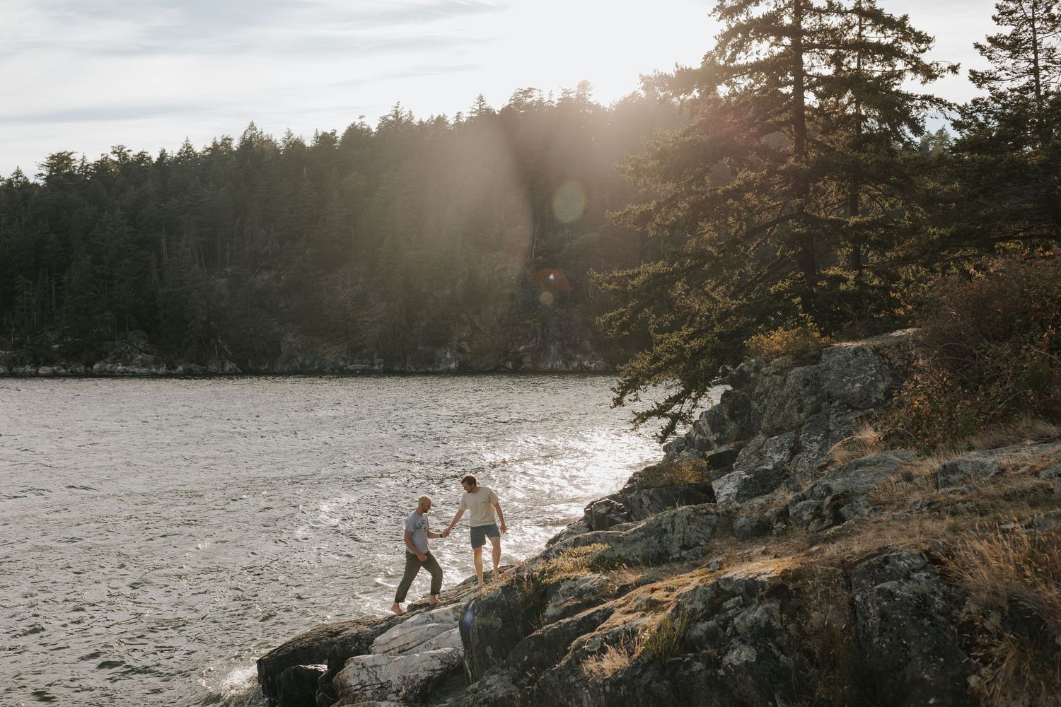Couple holding hands on rocky shoreline with sunlit forest and calm water in background.