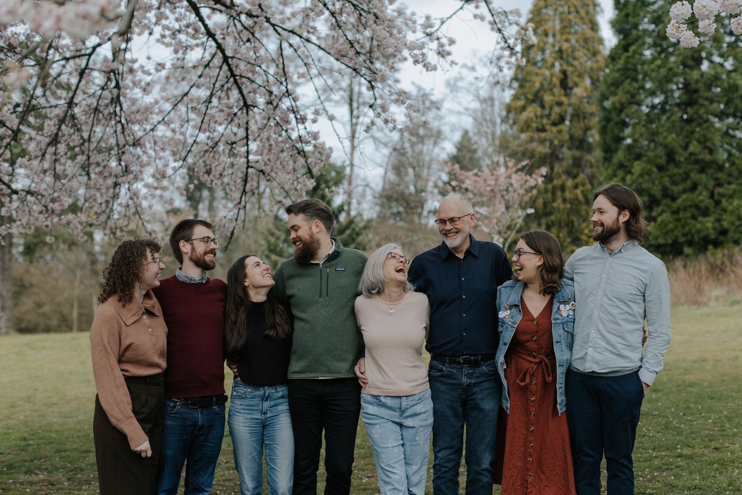 Seven family members smiling and embracing outdoors under blooming cherry blossom trees on a green lawn in spring.
