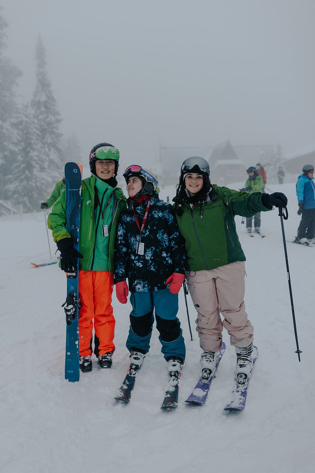 Three skiers in colorful winter gear pose together on a snowy slope with evergreen trees in the background.