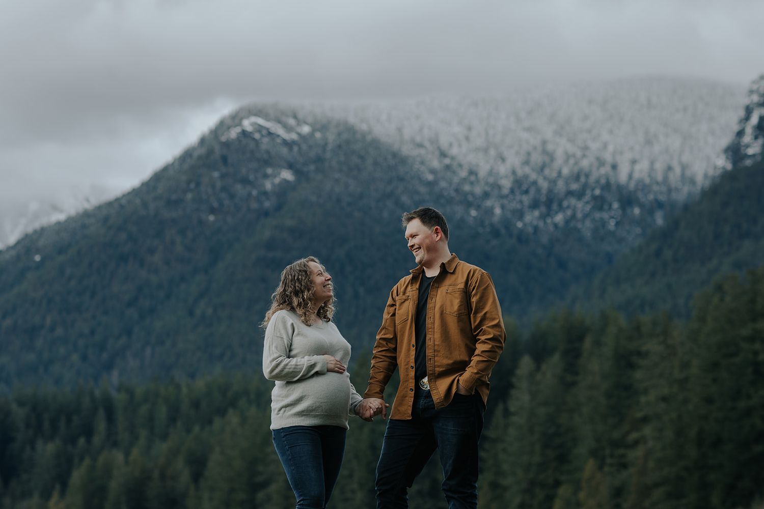 Couple standing together against backdrop of snow-dusted mountains and forest.