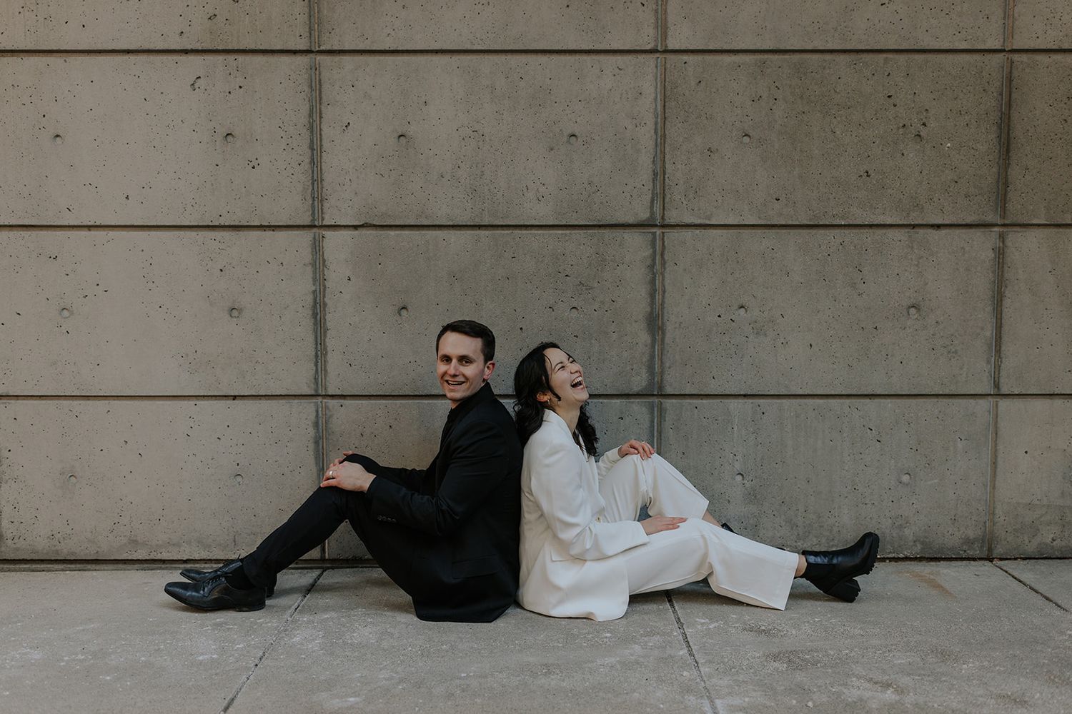A couple poses playfully against a concrete wall in contrasting black and white outfits.