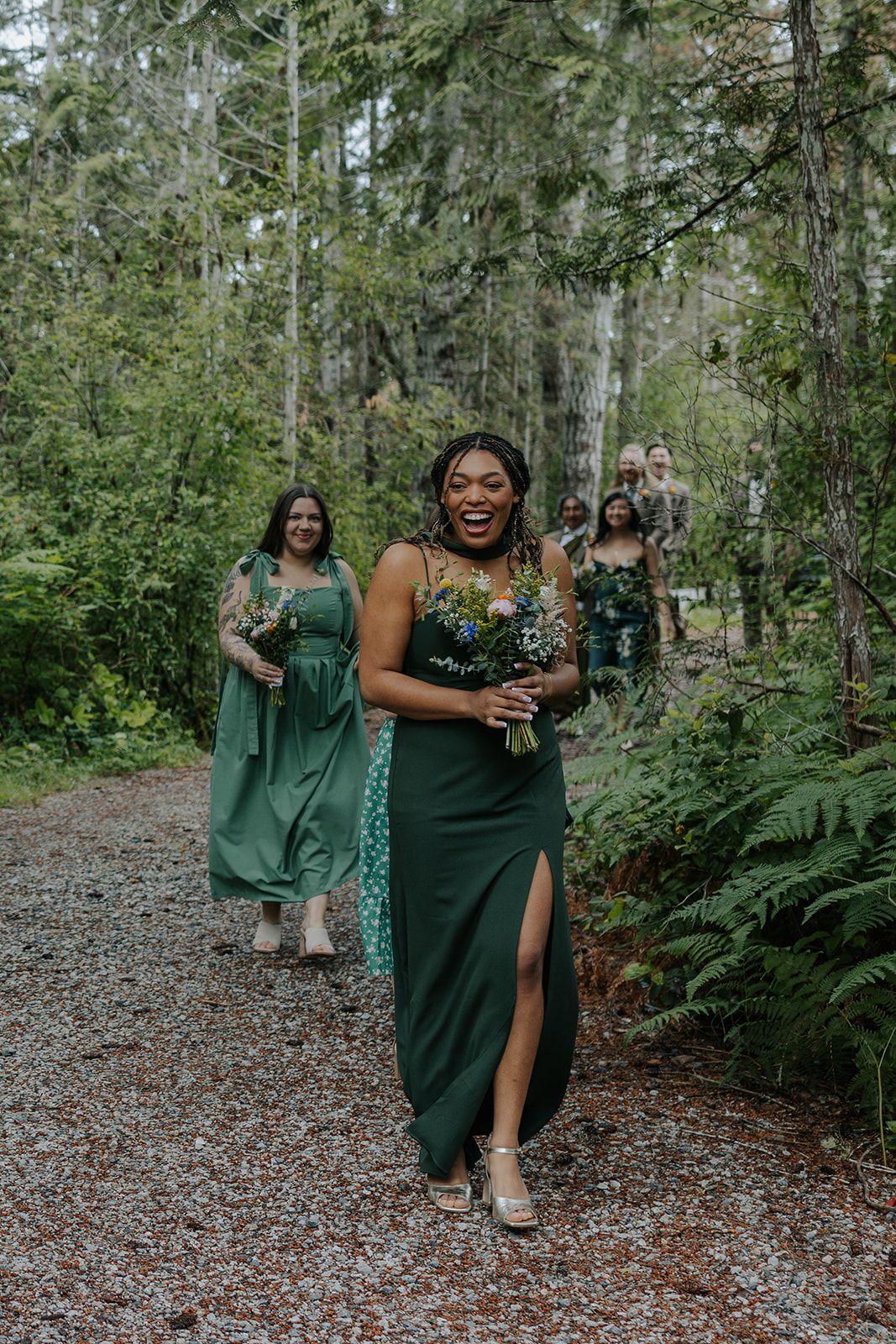 Wedding party members in emerald green dresses share joyful moments on a forest path.