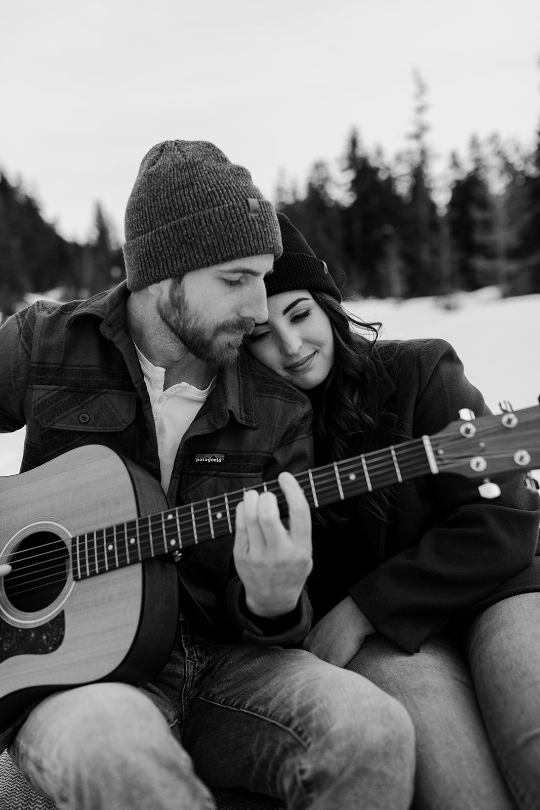 A couple shares an intimate moment while playing acoustic guitar in a snowy winter setting.