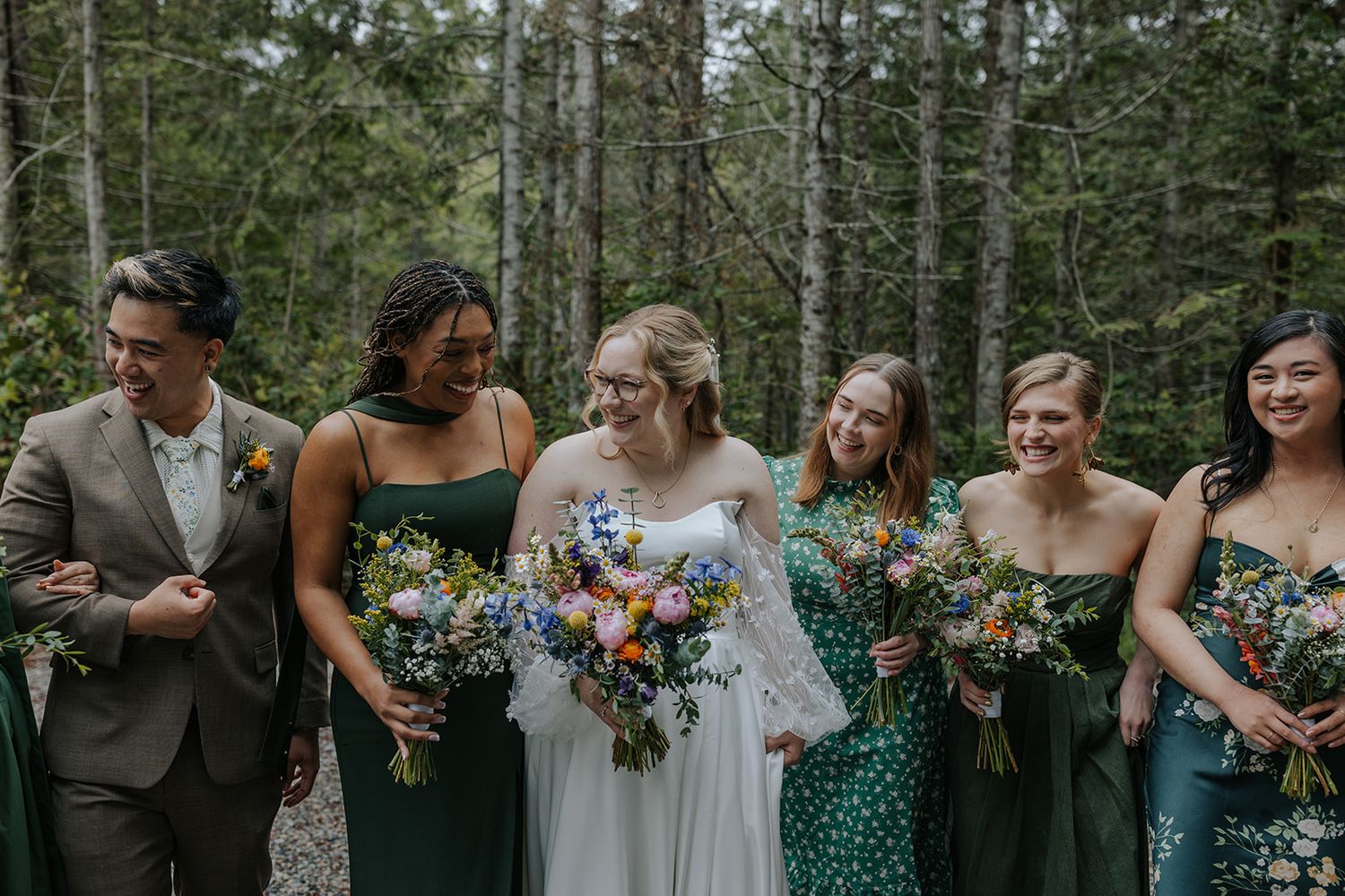 A wedding party poses outdoors in forest wearing green and white attire while holding colorful floral bouquets.