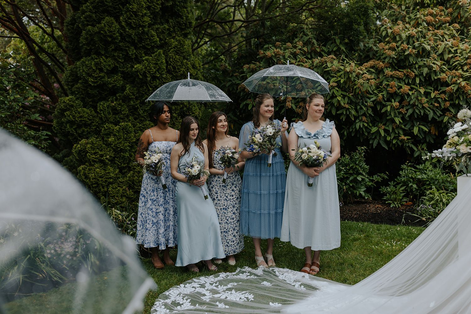 Wedding party holding clear umbrellas during rainy outdoor ceremony.