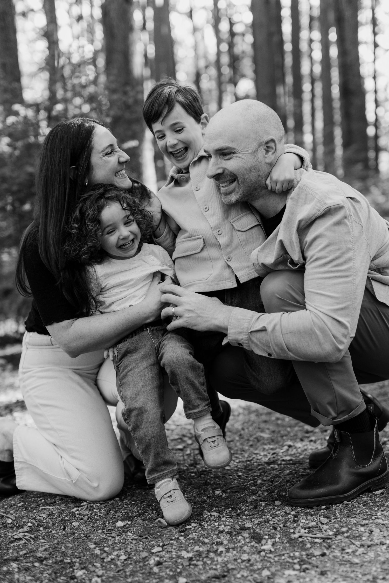 Black and white family portrait of parents and two young children laughing together in a forest setting.