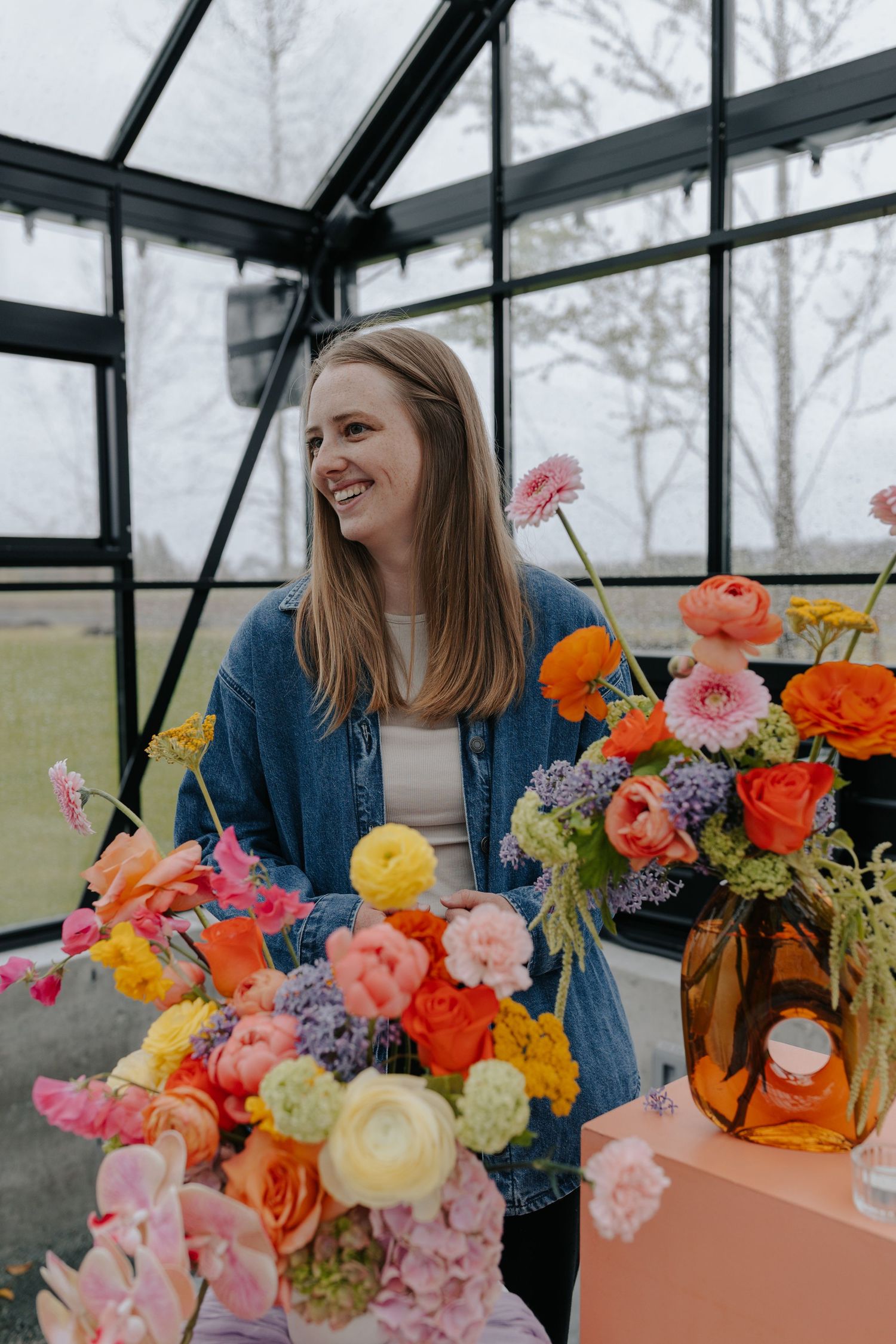 Smiling woman in denim jacket surrounded by colorful floral arrangements in a greenhouse setting.