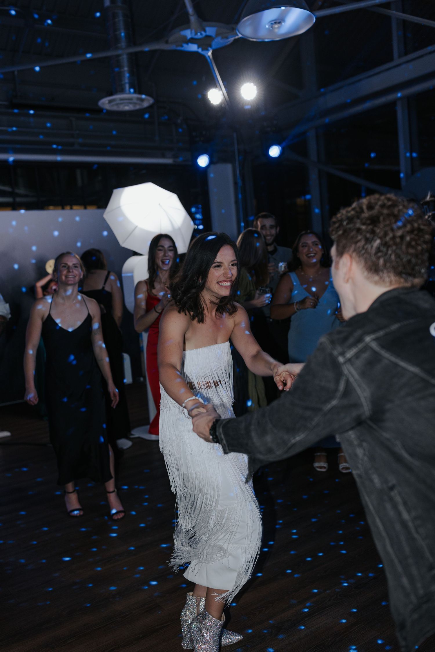 A woman in a white fringe dress dances at a wedding reception under blue lighting.