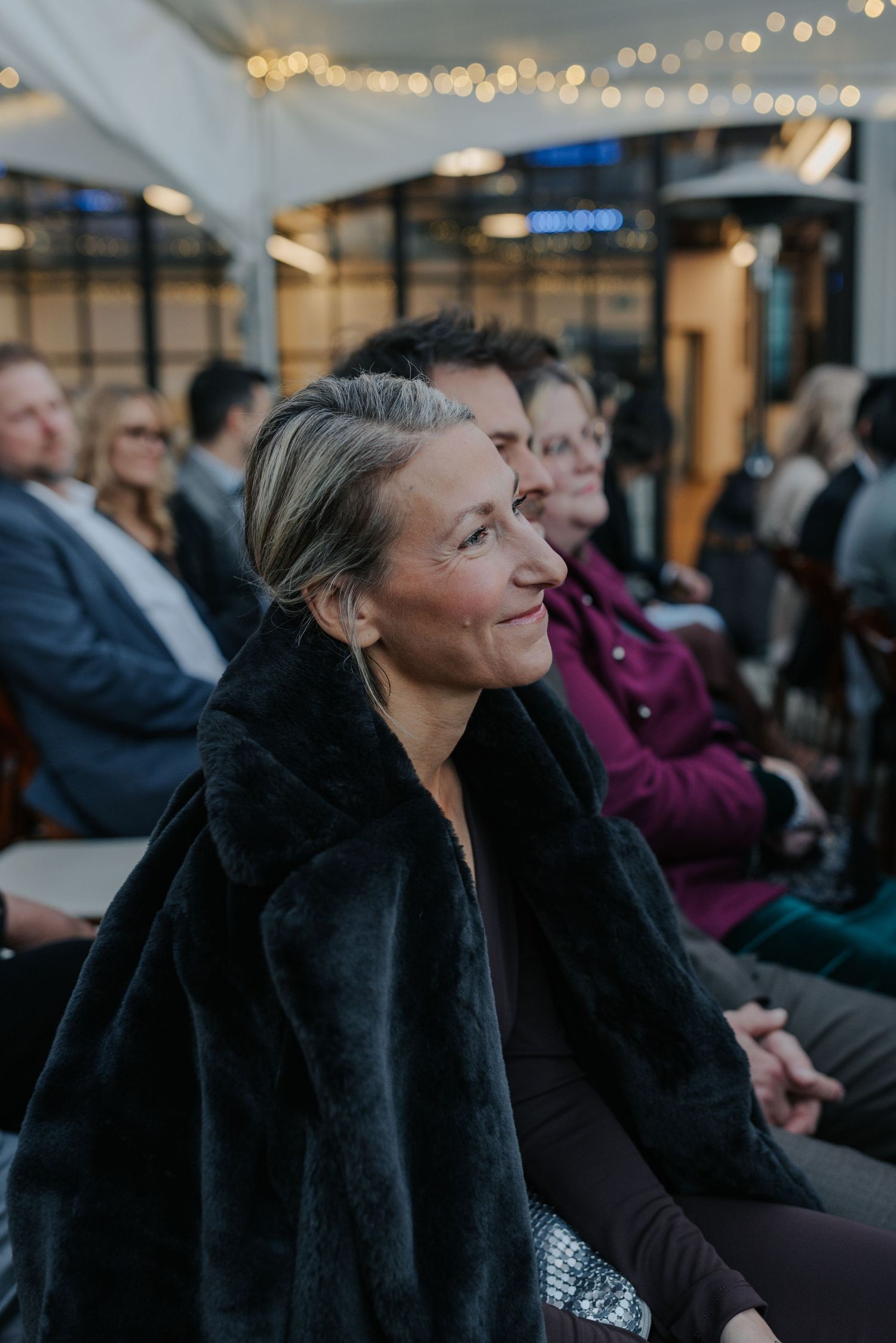 Guest in black coat enjoys outdoor wedding reception with string lights in background.