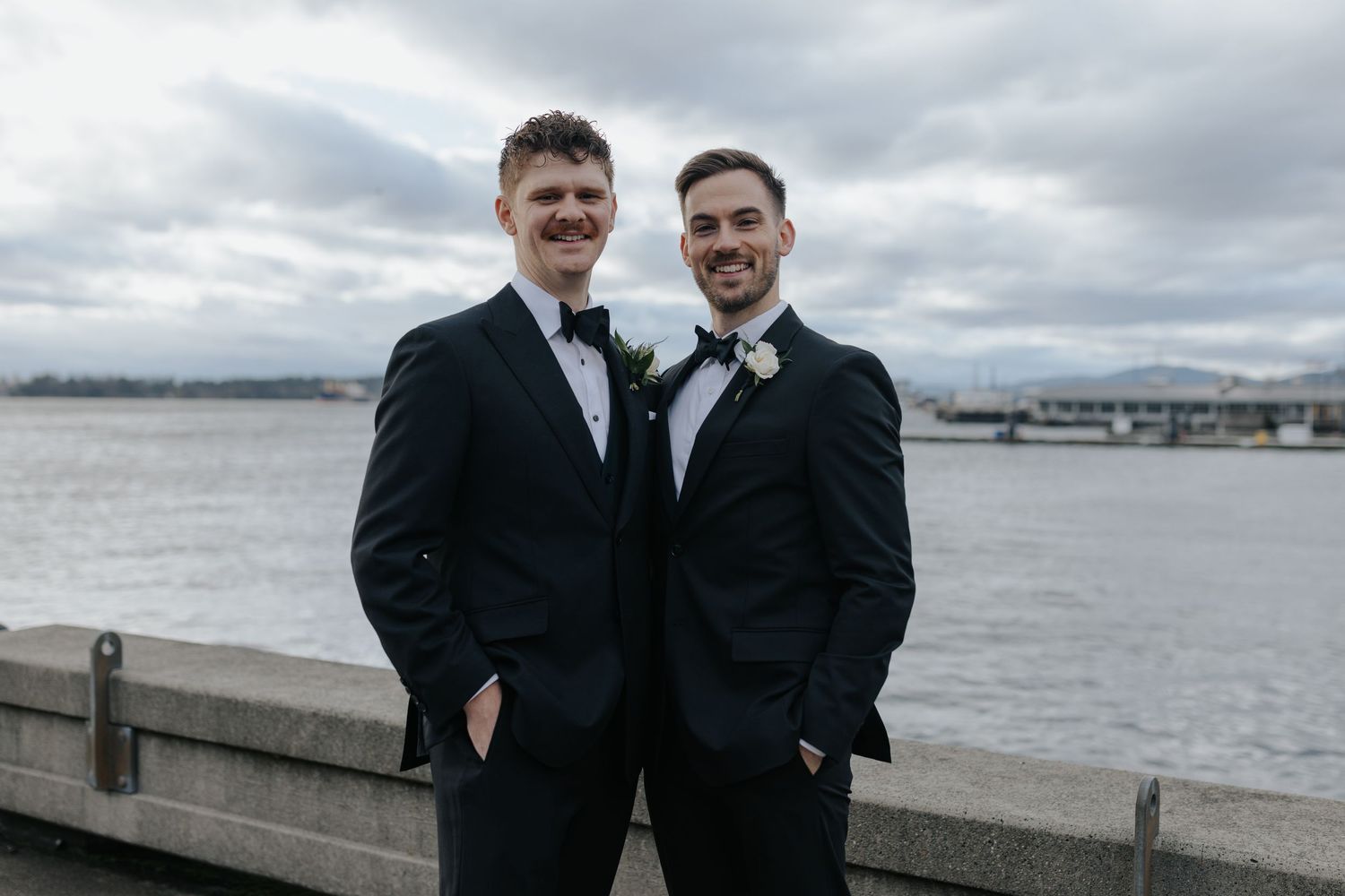 Two people in formal black suits stand together on waterfront with cloudy sky backdrop.