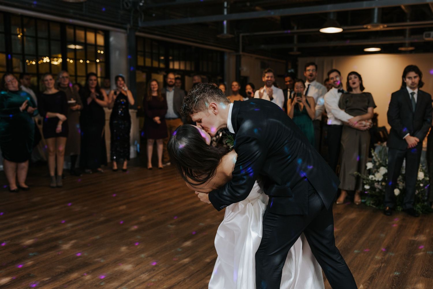 Wedding celebration on a wooden dance floor with guests dancing under ambient lighting.