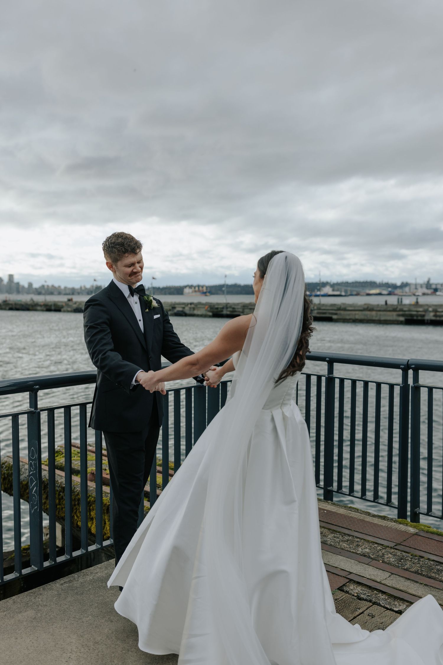 A couple shares a romantic moment on a waterfront deck during their wedding day under cloudy skies.