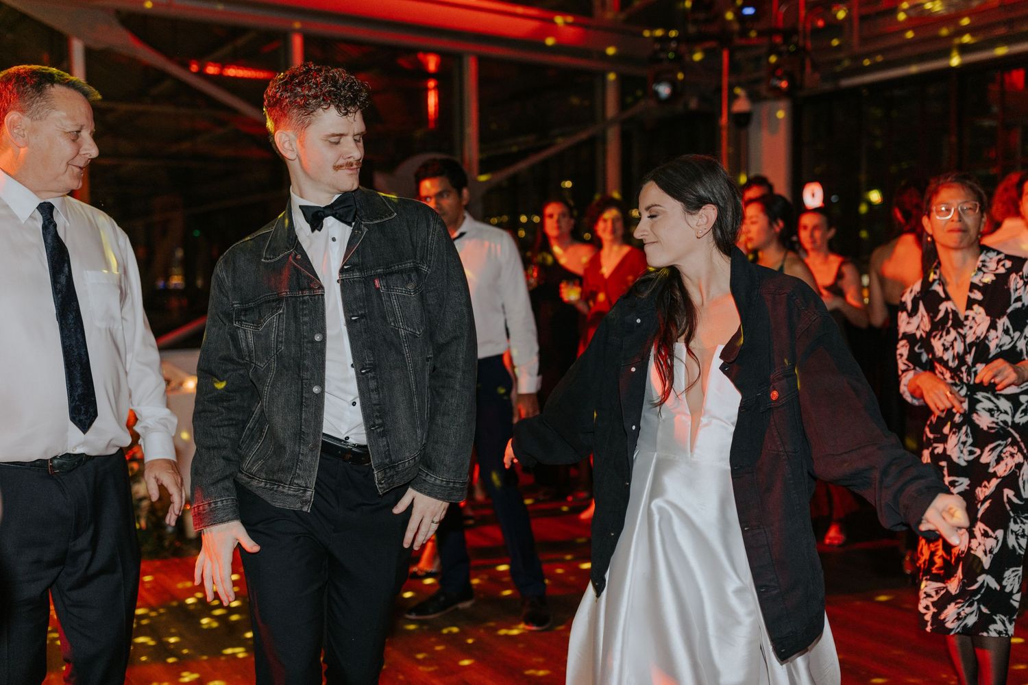 Wedding guests dance and celebrate on a dimly lit dance floor with red ambient lighting at an evening reception.