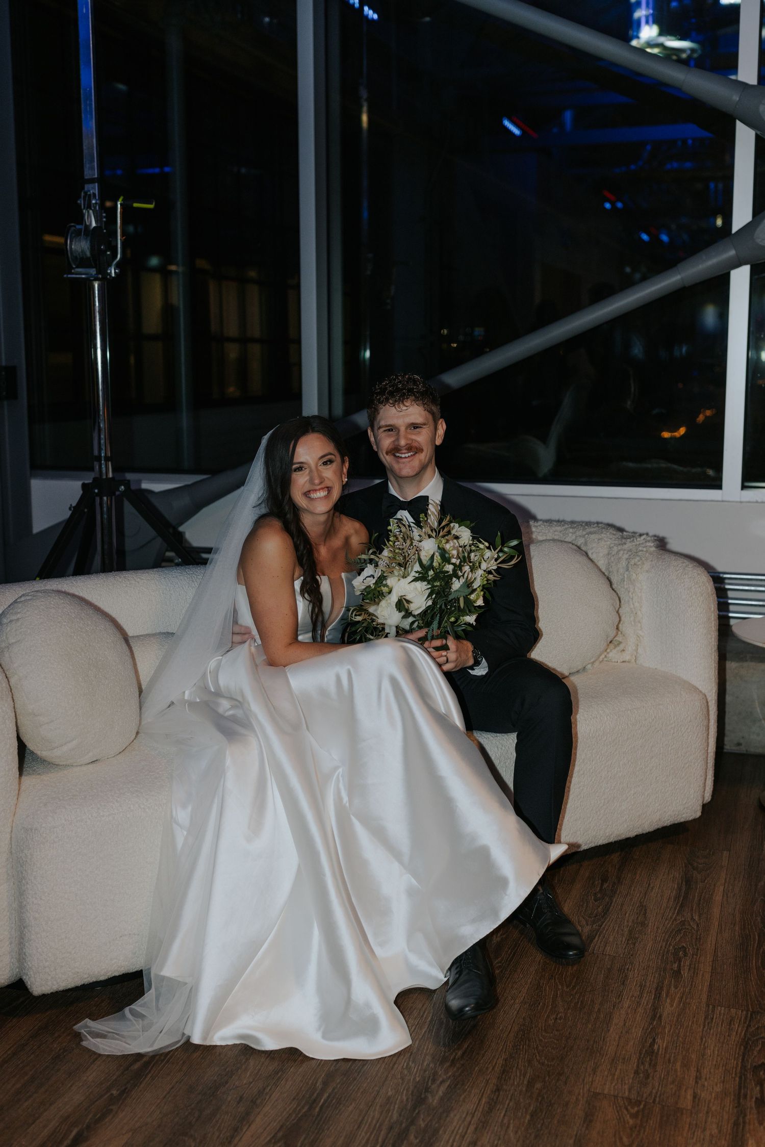 Newlyweds sit together on a cream colored couch, with the bride holding a white floral bouquet.