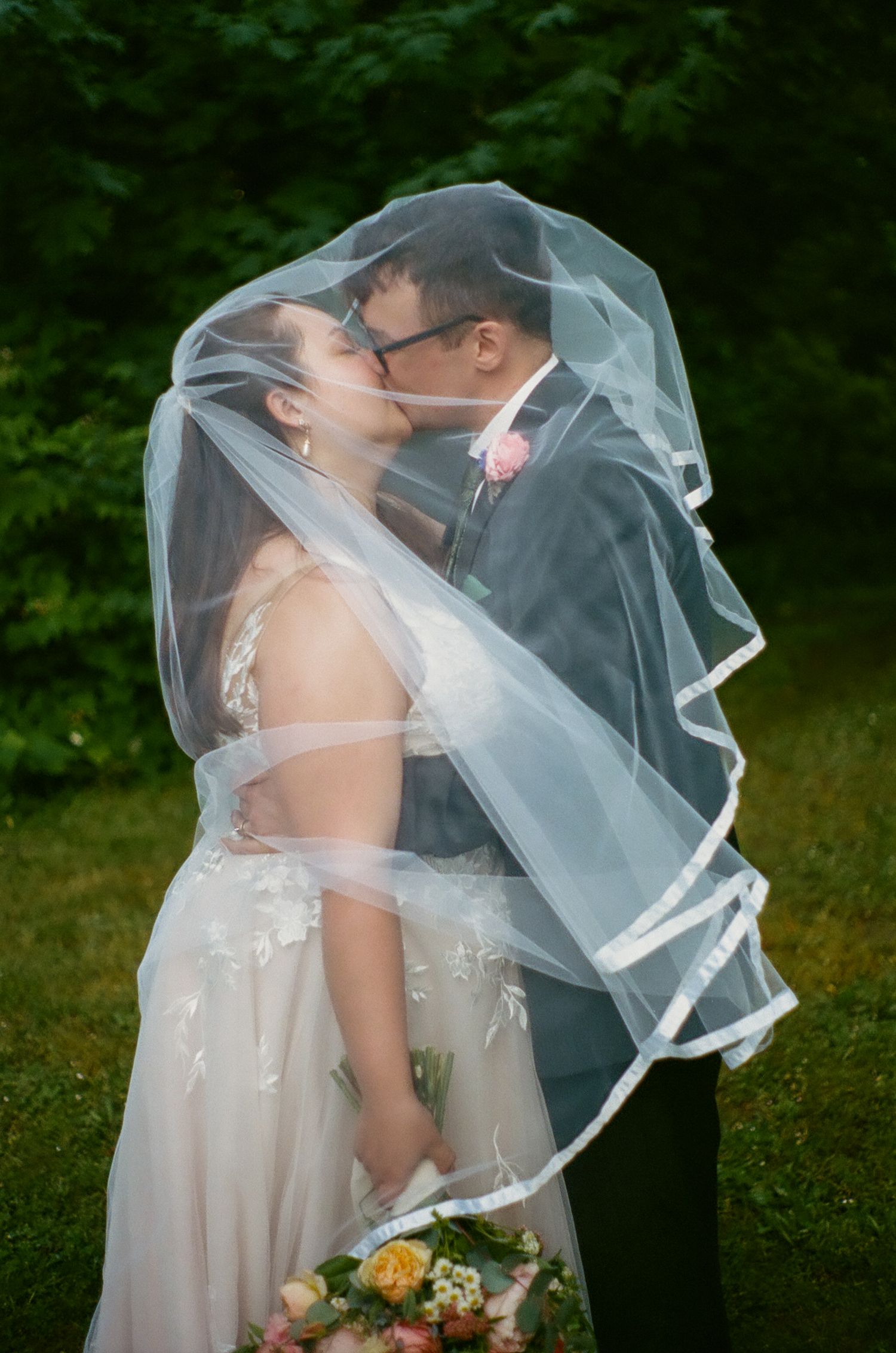 A romantic wedding couple shares a kiss under a flowing veil while holding a peach and white floral bouquet outdoors.